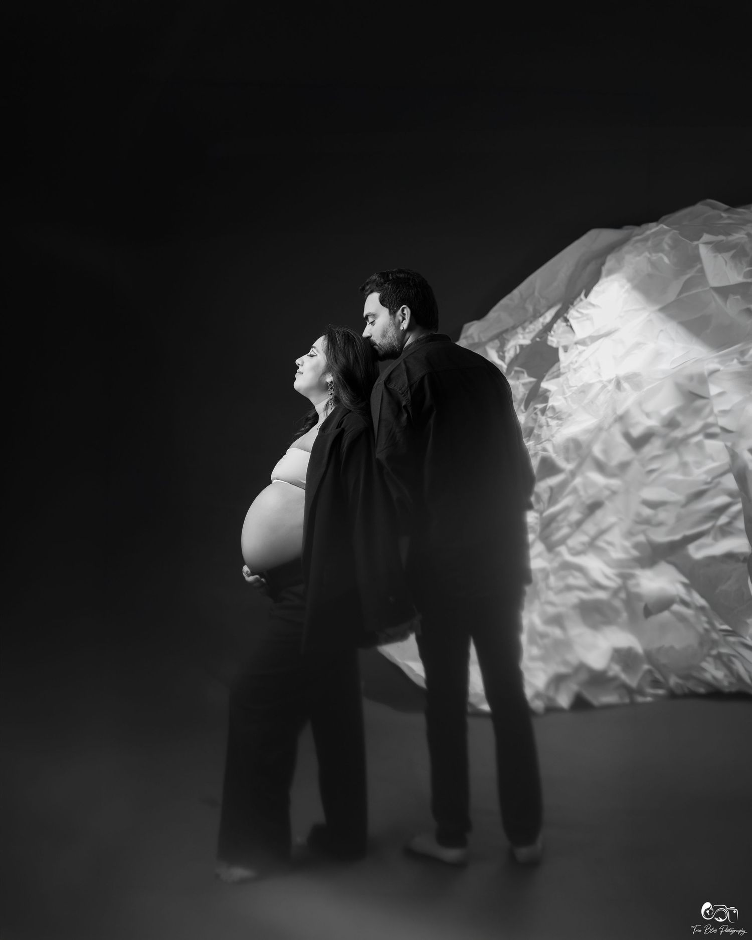 Black and white maternity photo of pregnant woman and partner posing against crinkled paper backdrop in studio.