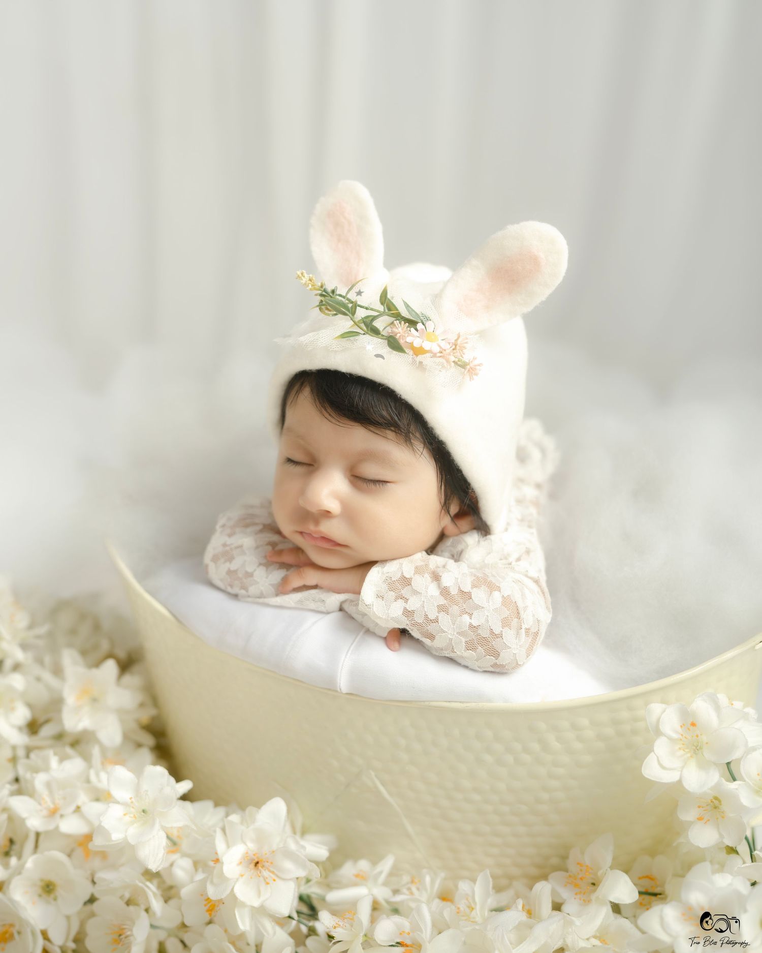 Sleeping newborn baby in bunny ear bonnet with flowers, posed in cream bowl surrounded by white blossoms.