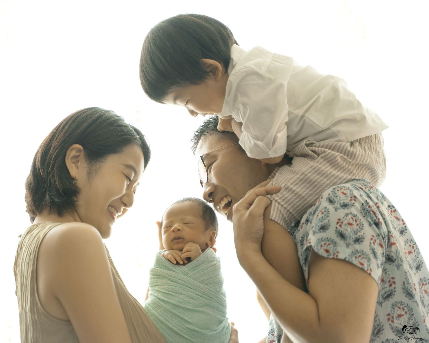 A joyful Asian family with a toddler on dad's shoulders and mom cradling a swaddled newborn baby.
