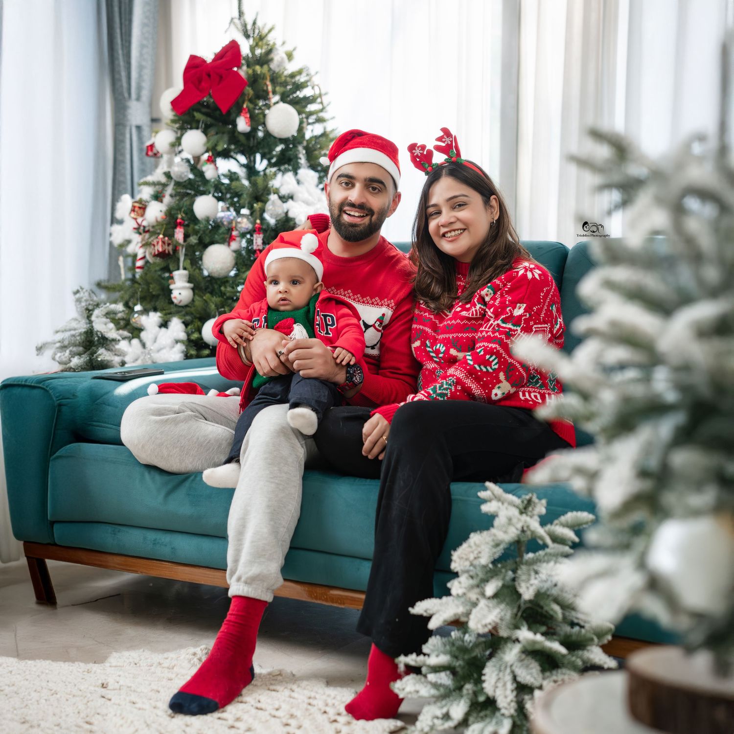Family of three in matching red Christmas sweaters and Santa hats sitting on a teal sofa near a decorated Christmas tree.