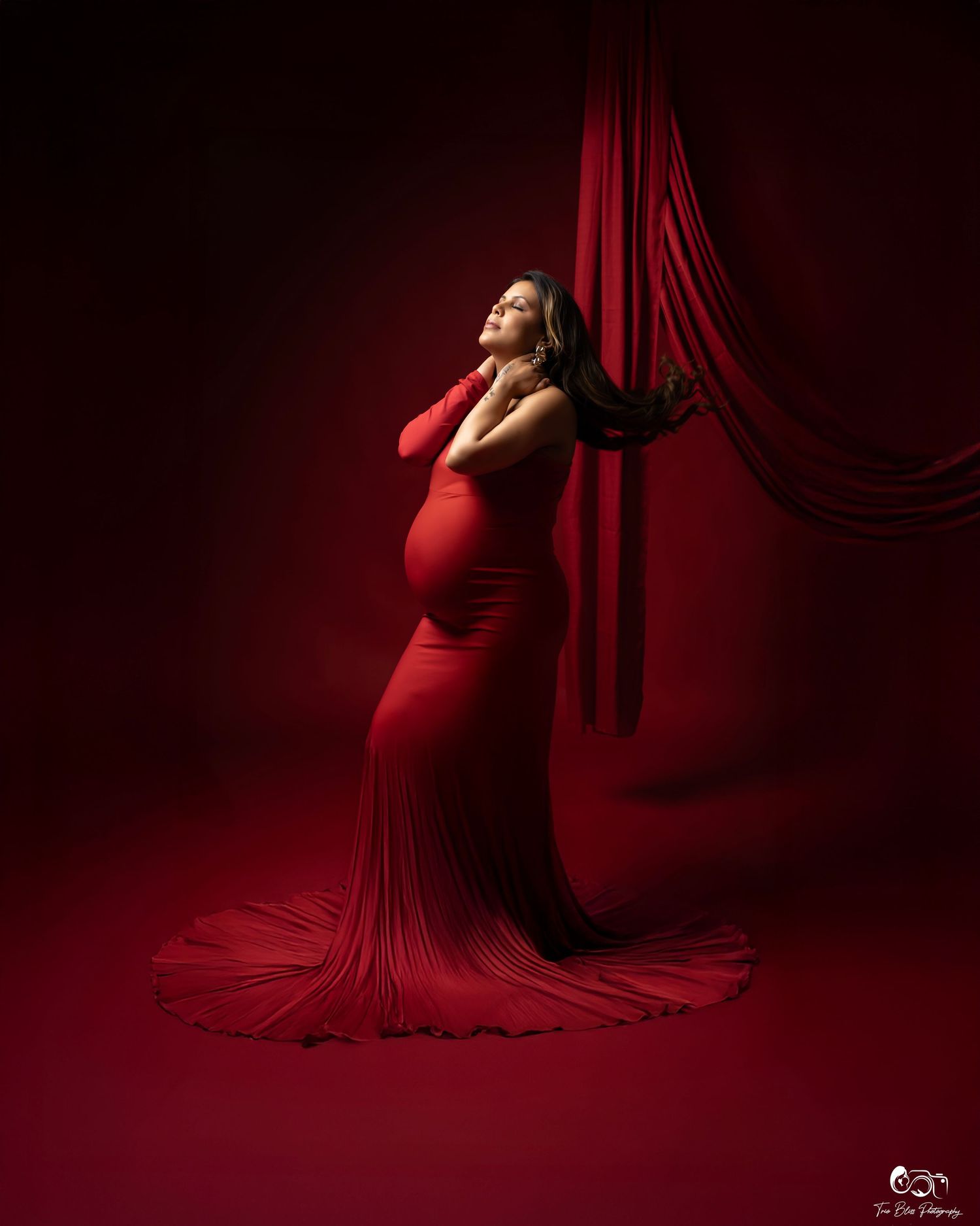 Maternity portrait in dark red tones showing silhouette against flowing fabric in dramatic studio lighting.