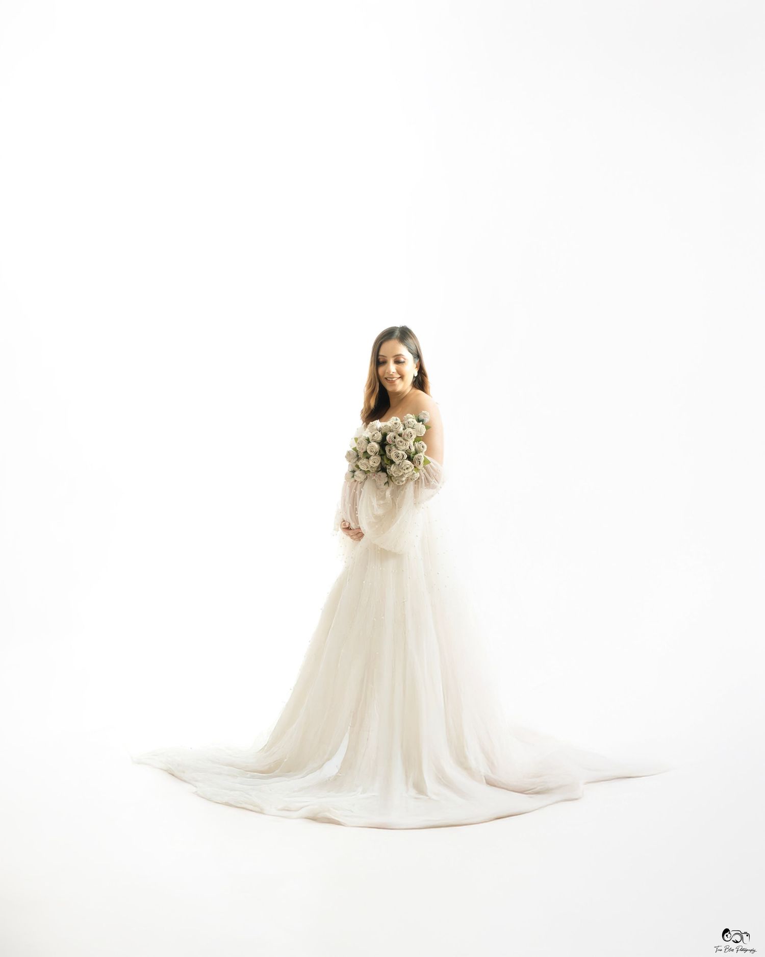 Bride in elegant white off-shoulder gown with long train holds bouquet, posing in bright studio.