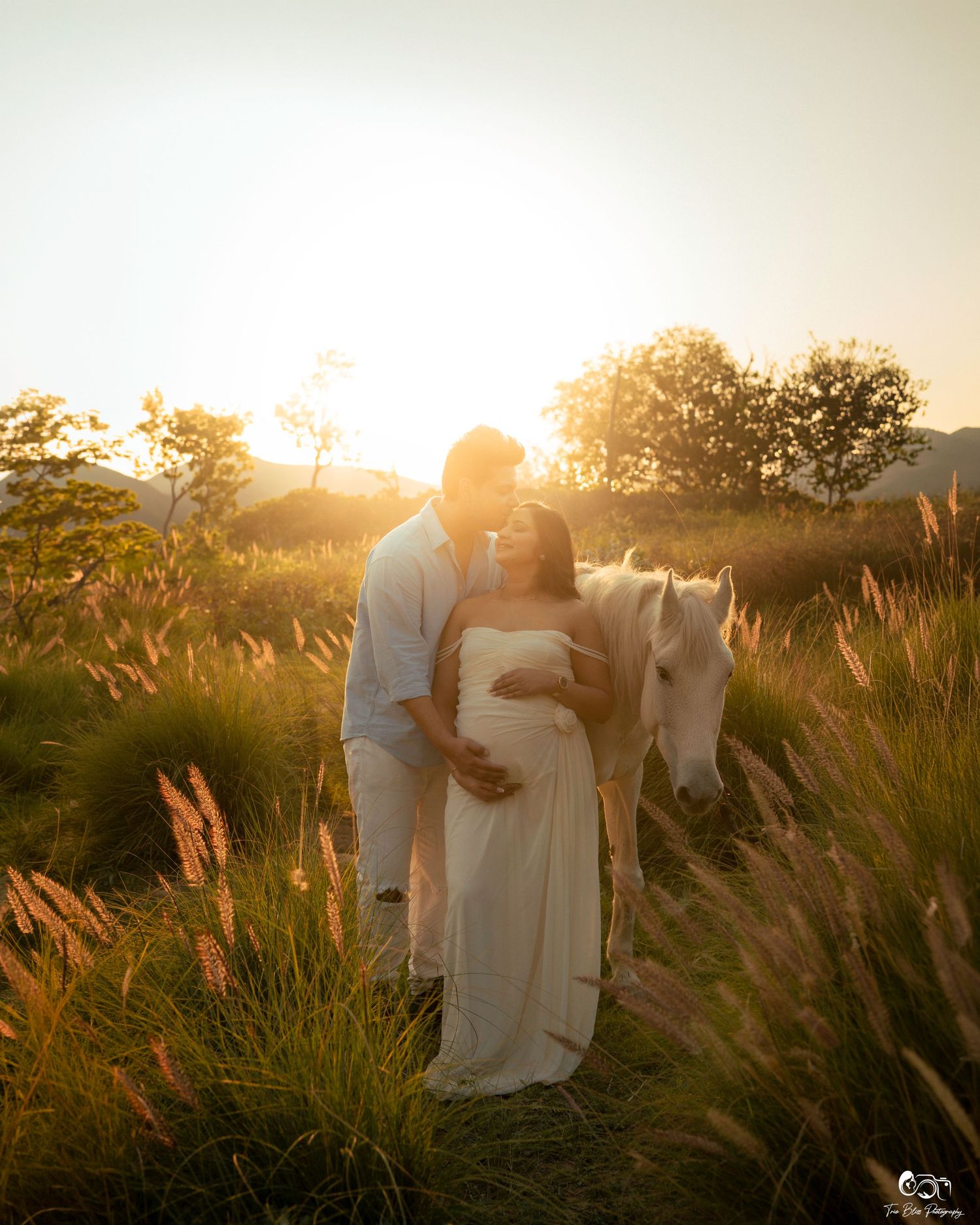 Couple maternity photoshoot at golden sunset in tall grass field with white horse, woman in flowing white gown.