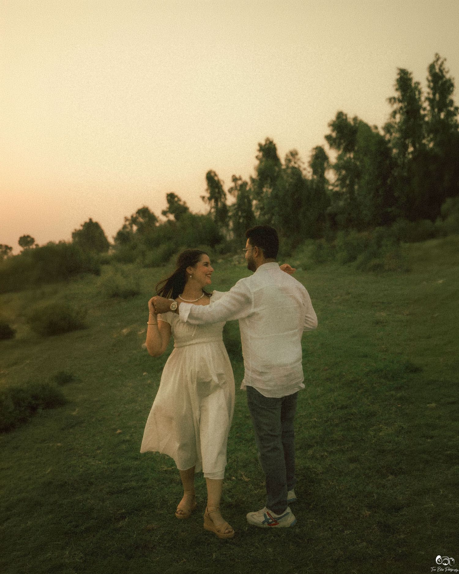 Couple dancing together on a green hillside at golden hour sunset, woman in white dress, man in white shirt and jeans.