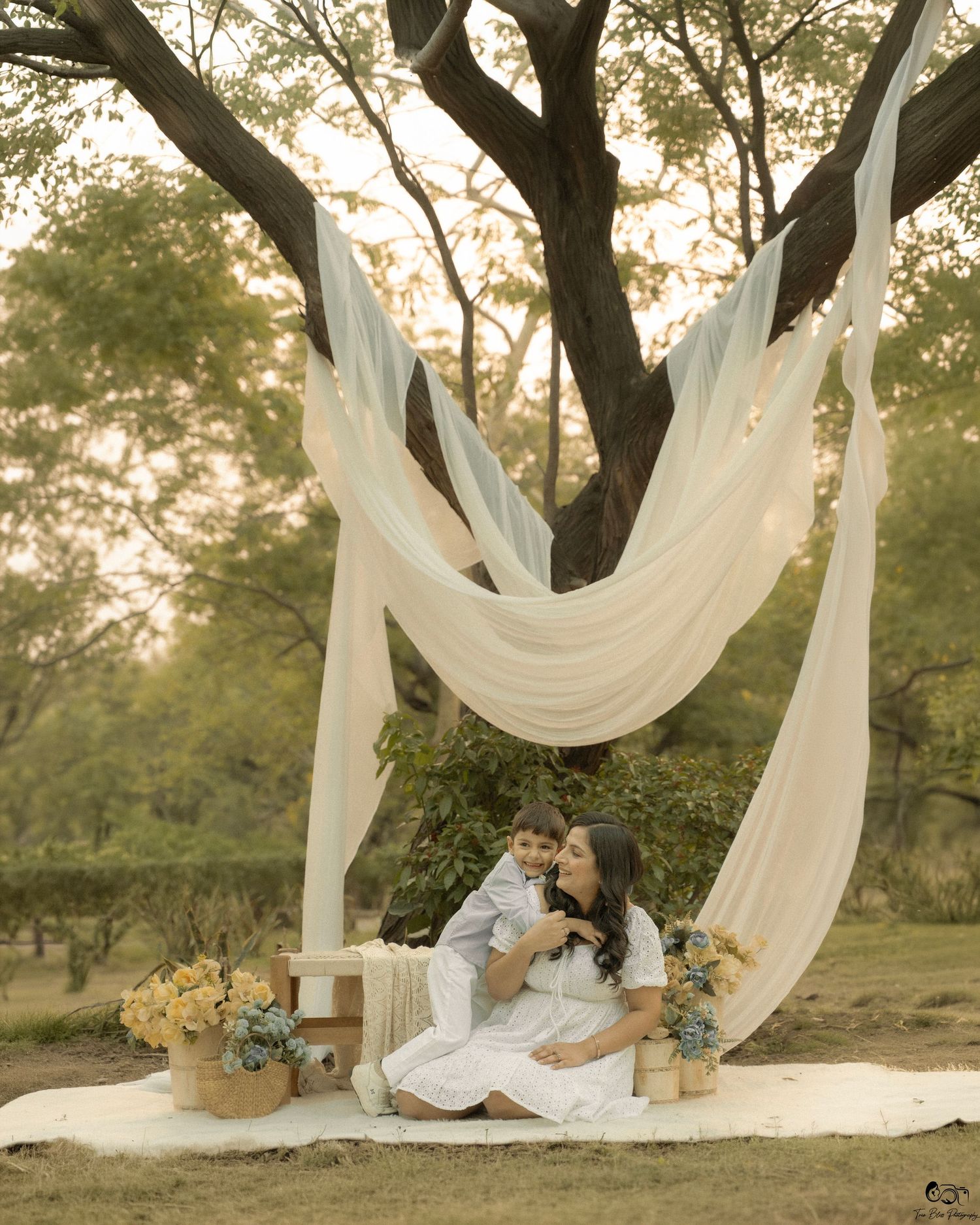 A mother and child sit together under a tree with white draping, surrounded by yellow flowers in a golden hour outdoor setting.