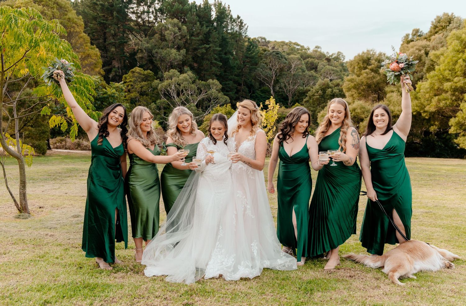 Wedding party in emerald green dresses poses joyfully on grassy lawn surrounded by autumn trees.