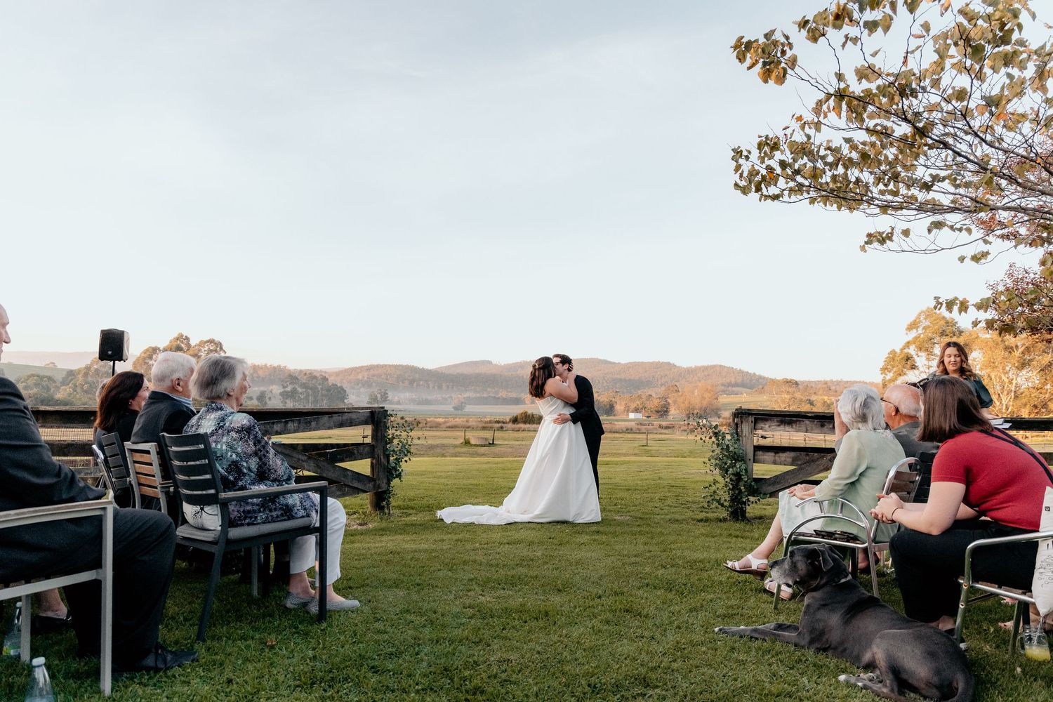 Outdoor wedding ceremony with guests seated on a grassy lawn overlooking scenic rolling hills at sunset.