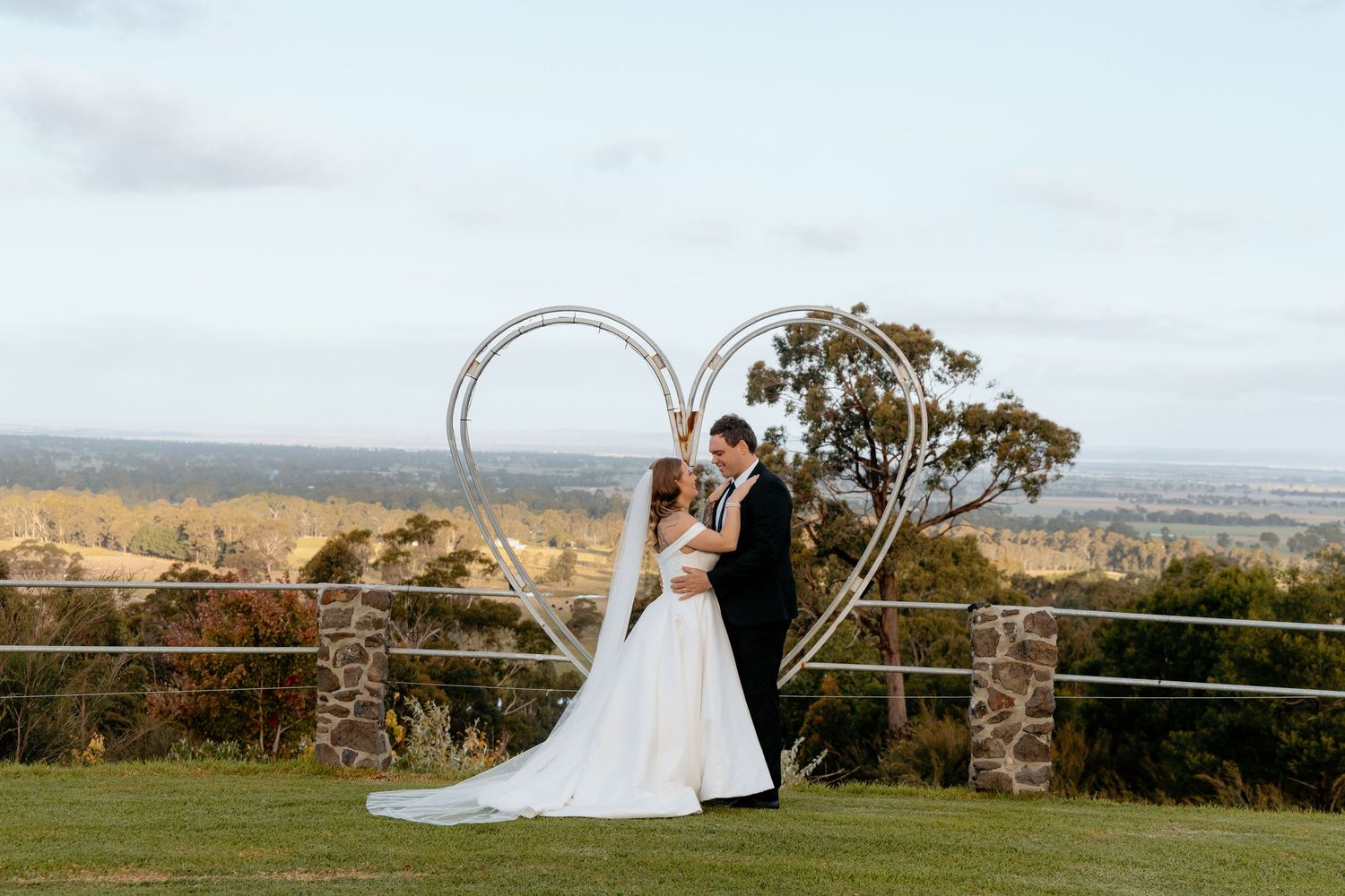 Bride and groom kissing under a heart-shaped arch with scenic countryside views at an outdoor wedding.