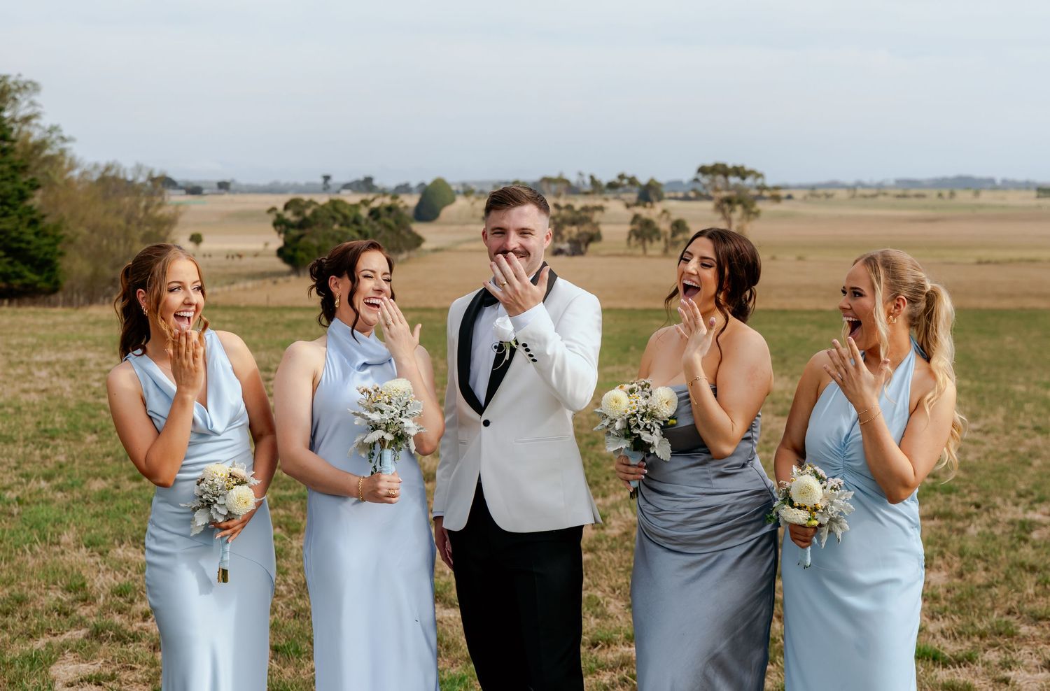Groom in white tuxedo laughing with four bridesmaids in light blue dresses holding bouquets in an open field.