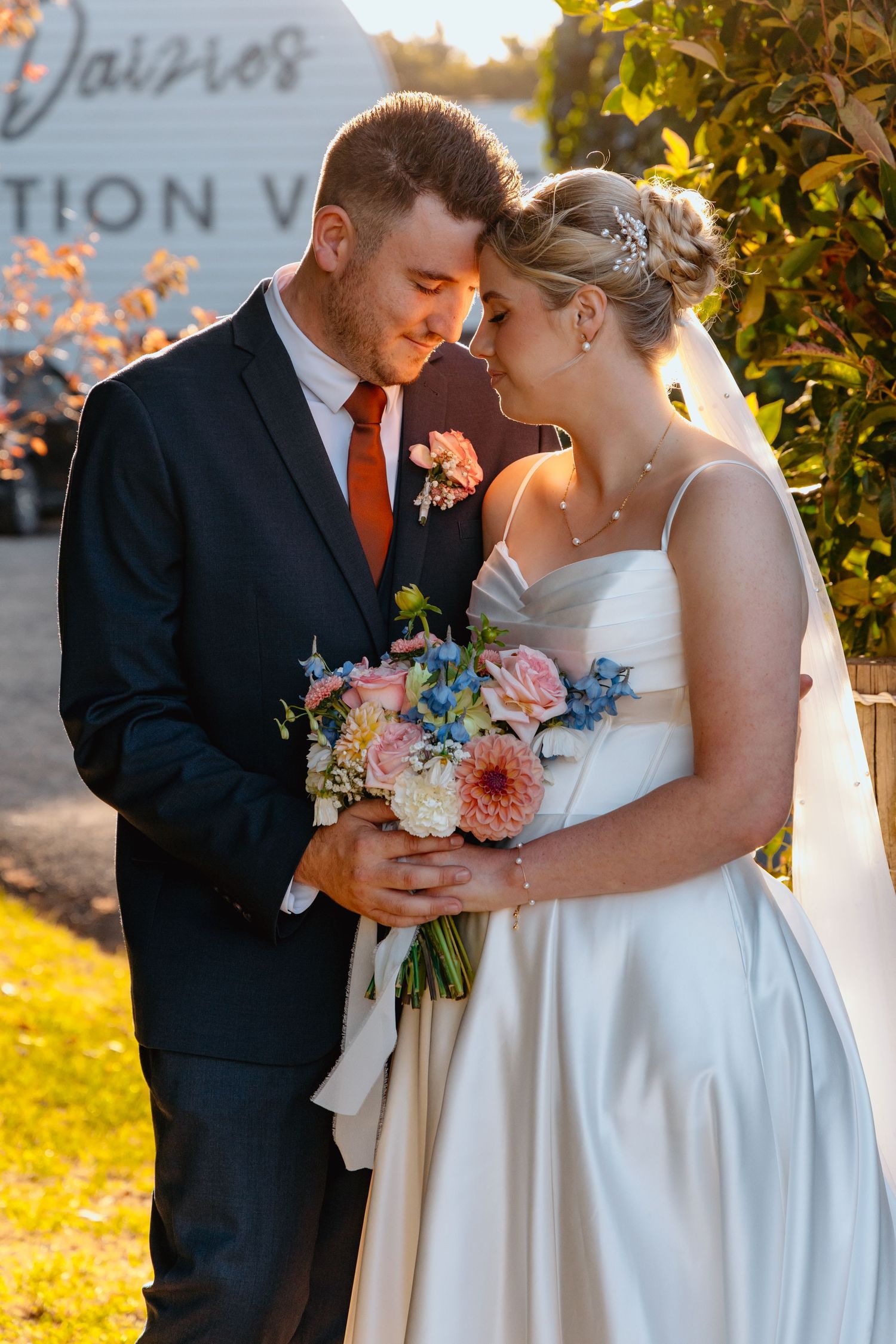 Bride and groom share a romantic forehead touch holding a colorful pink and blue bouquet at an outdoor wedding venue.