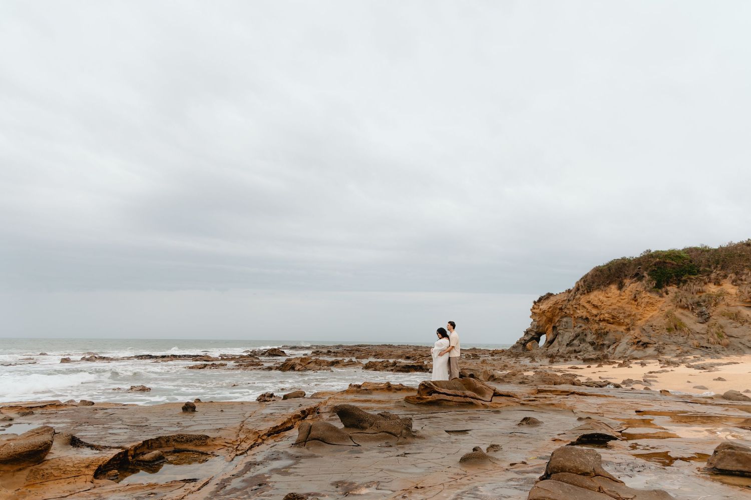 A figure in white stands on rocky coastal cliffs overlooking a misty ocean landscape at sunset.