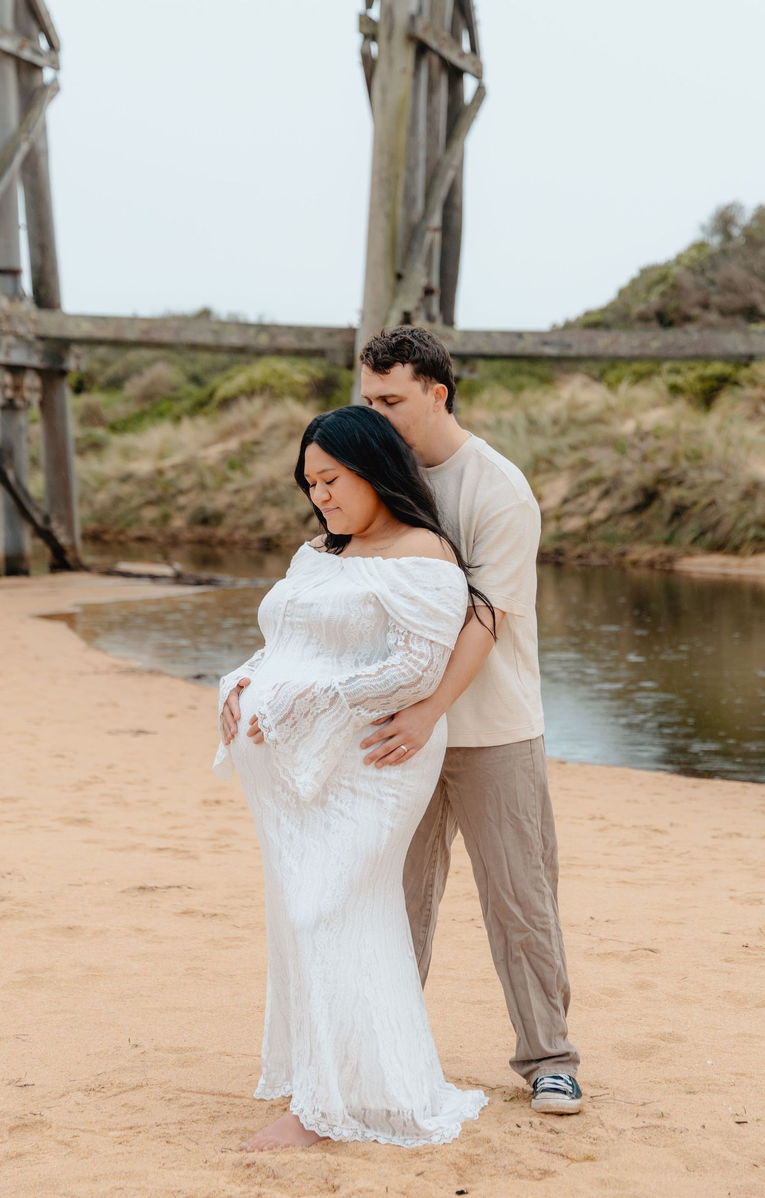 A couple in casual attire embraces tenderly on a sandy beach near a tranquil waterfront.