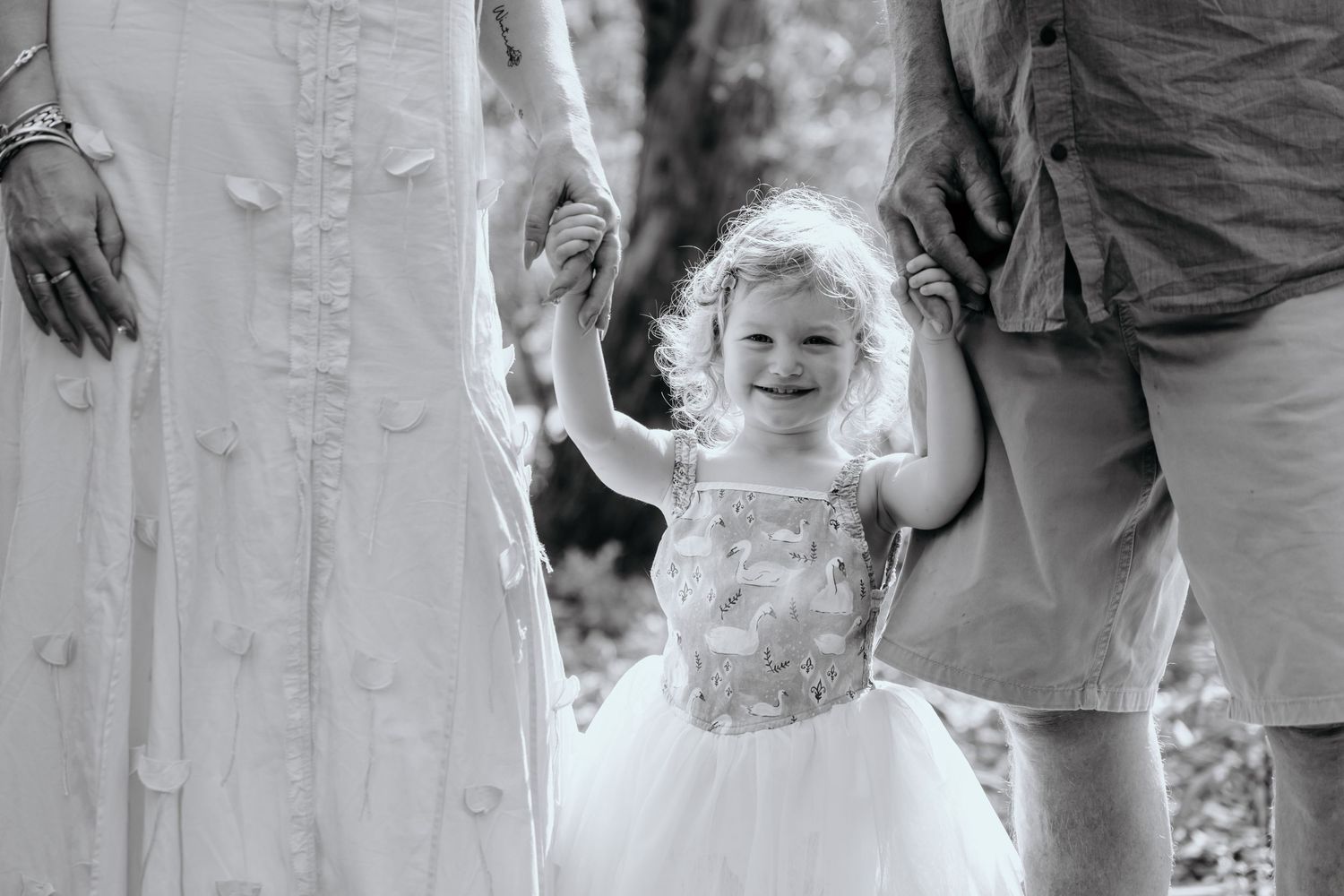 Black and white portrait of joyful child in tulle dress holding hands with parents.