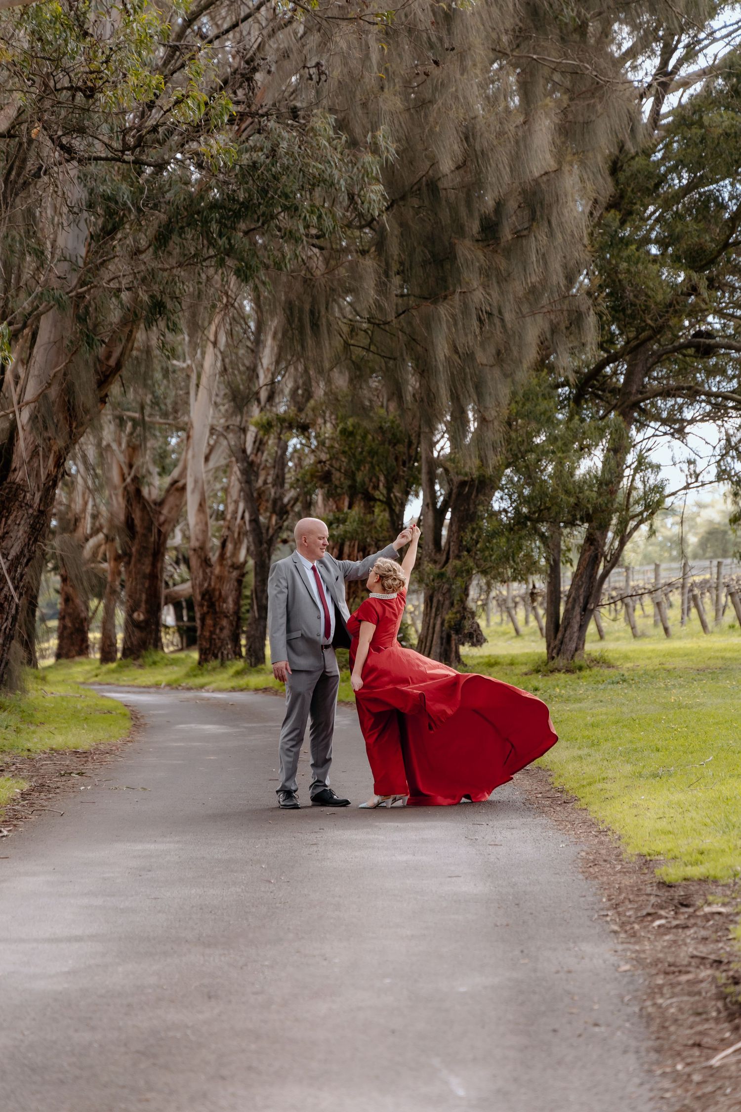 A romantic couple poses for photos along a tree-lined path, with the woman wearing a flowing red dress.