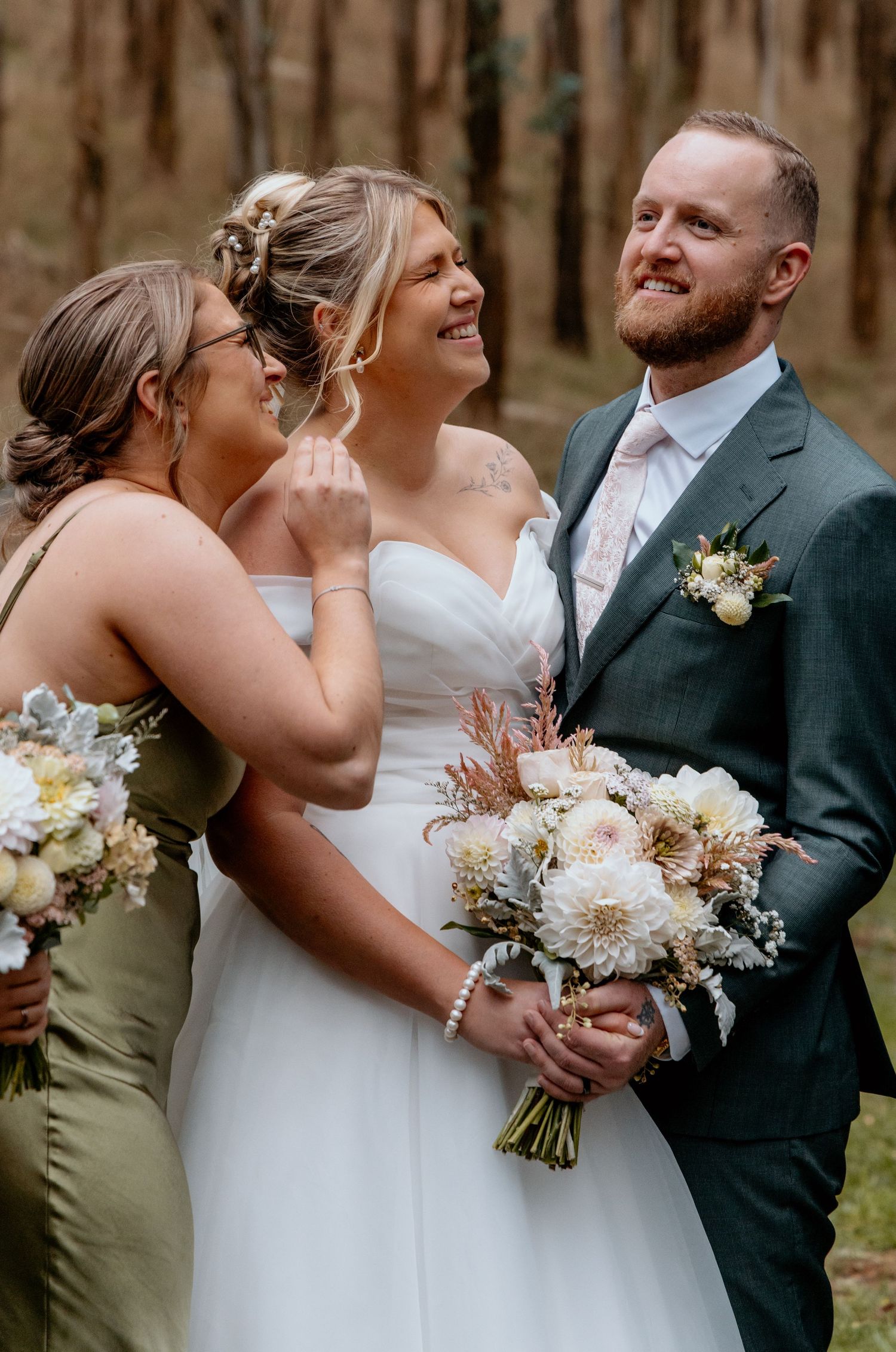 Bride in white gown laughs with bridesmaid in olive dress as groom in teal suit smiles, forest wedding backdrop.