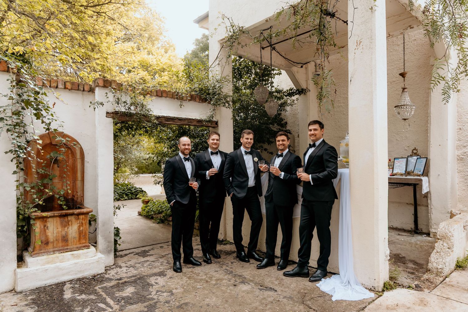 Groom and groomsmen in black tuxedos smiling together outside a white stucco venue with lush greenery.