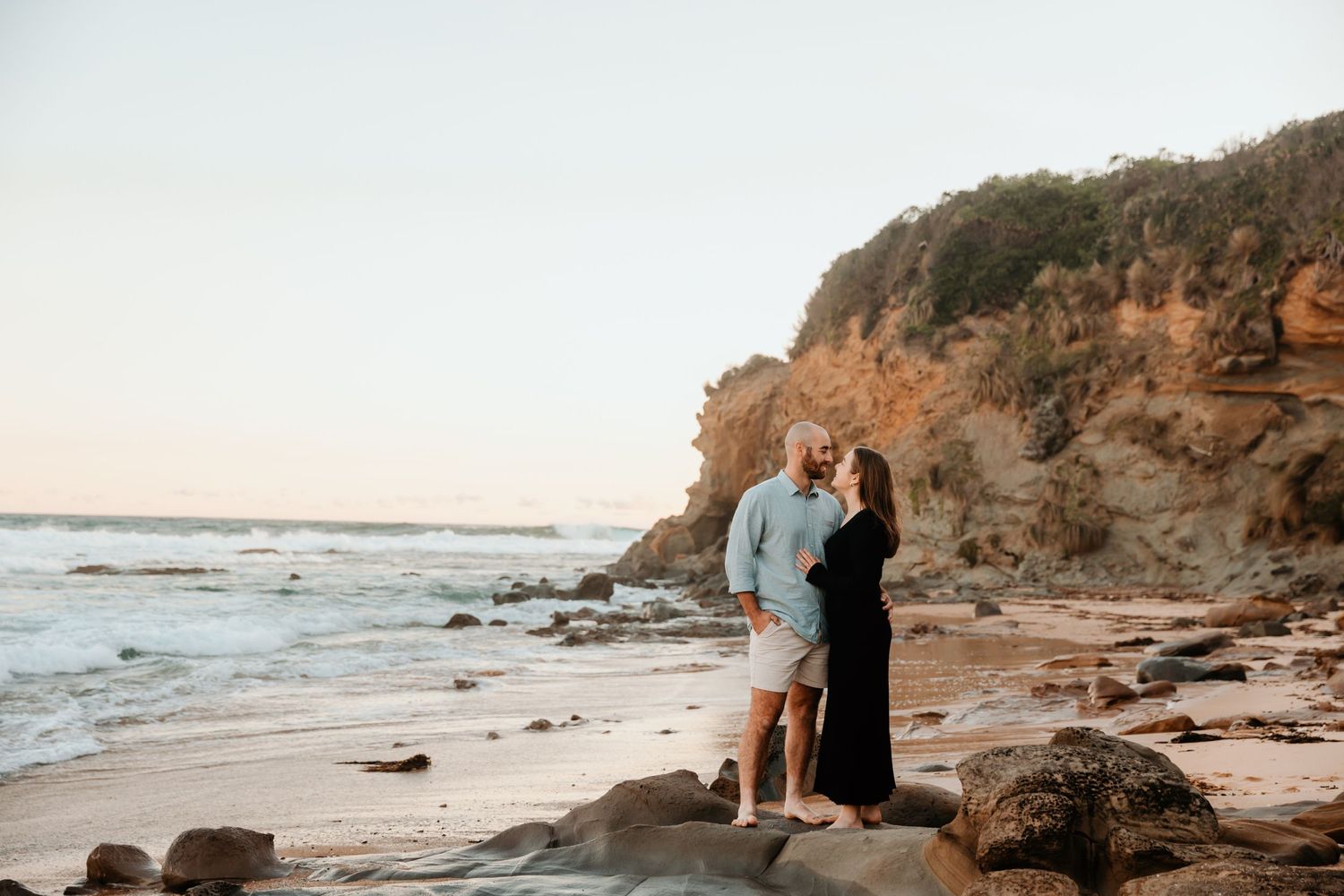 Couple stands on rocky beach with dramatic coastal cliffs during sunset.