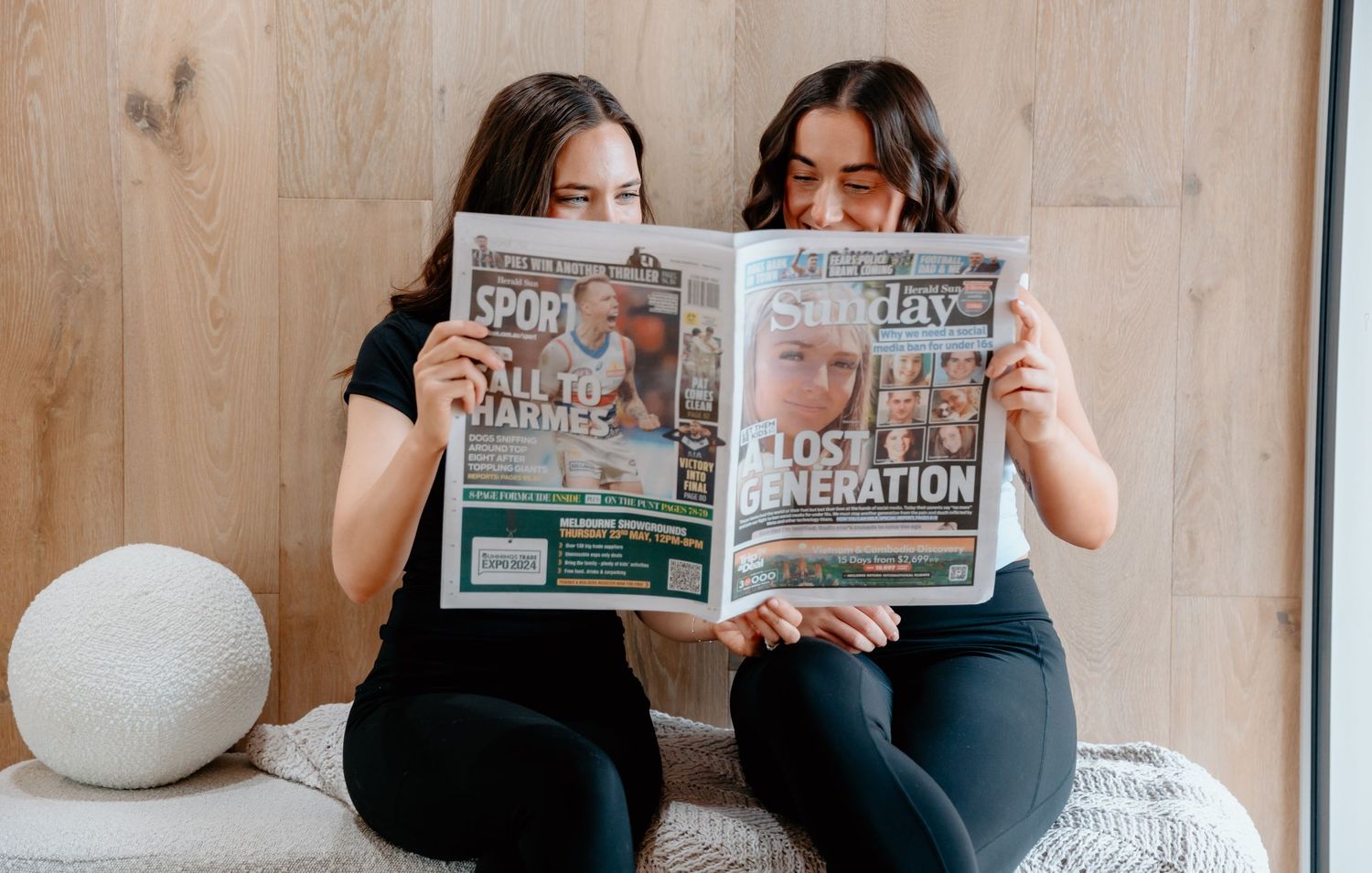 Two people on couch reading newspapers together in bright room with wooden floors.