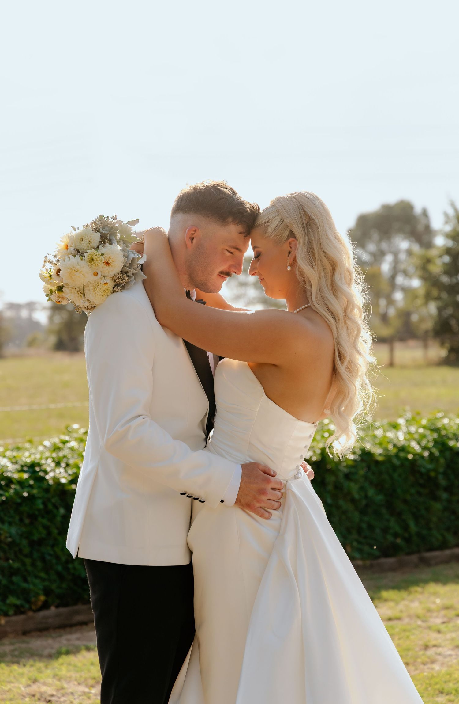 Bride and groom in white wedding attire embrace forehead-to-forehead outdoors, holding a white floral bouquet.