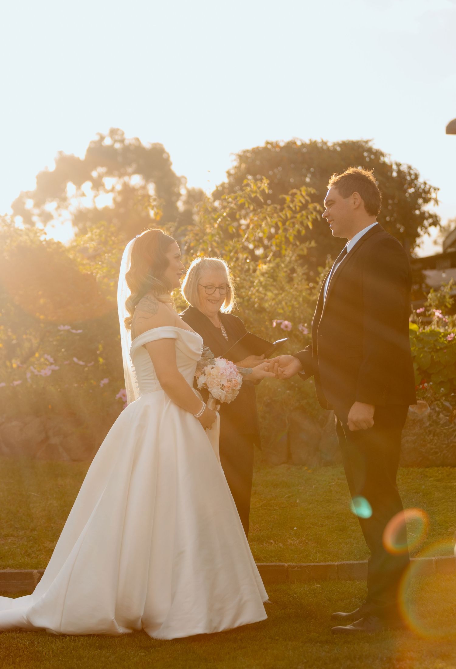 Bride and groom exchange vows outdoors at golden hour with officiant, surrounded by lush greenery and warm sunlight.