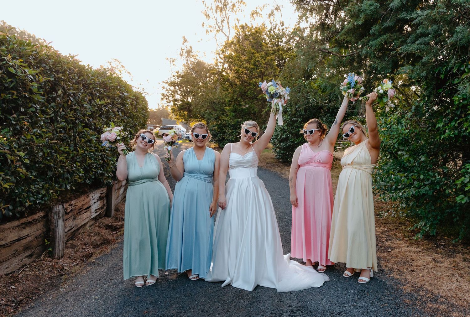 Bride and bridesmaids in rainbow pastel gowns and sunglasses raising bouquets outdoors on a sunny wedding day.
