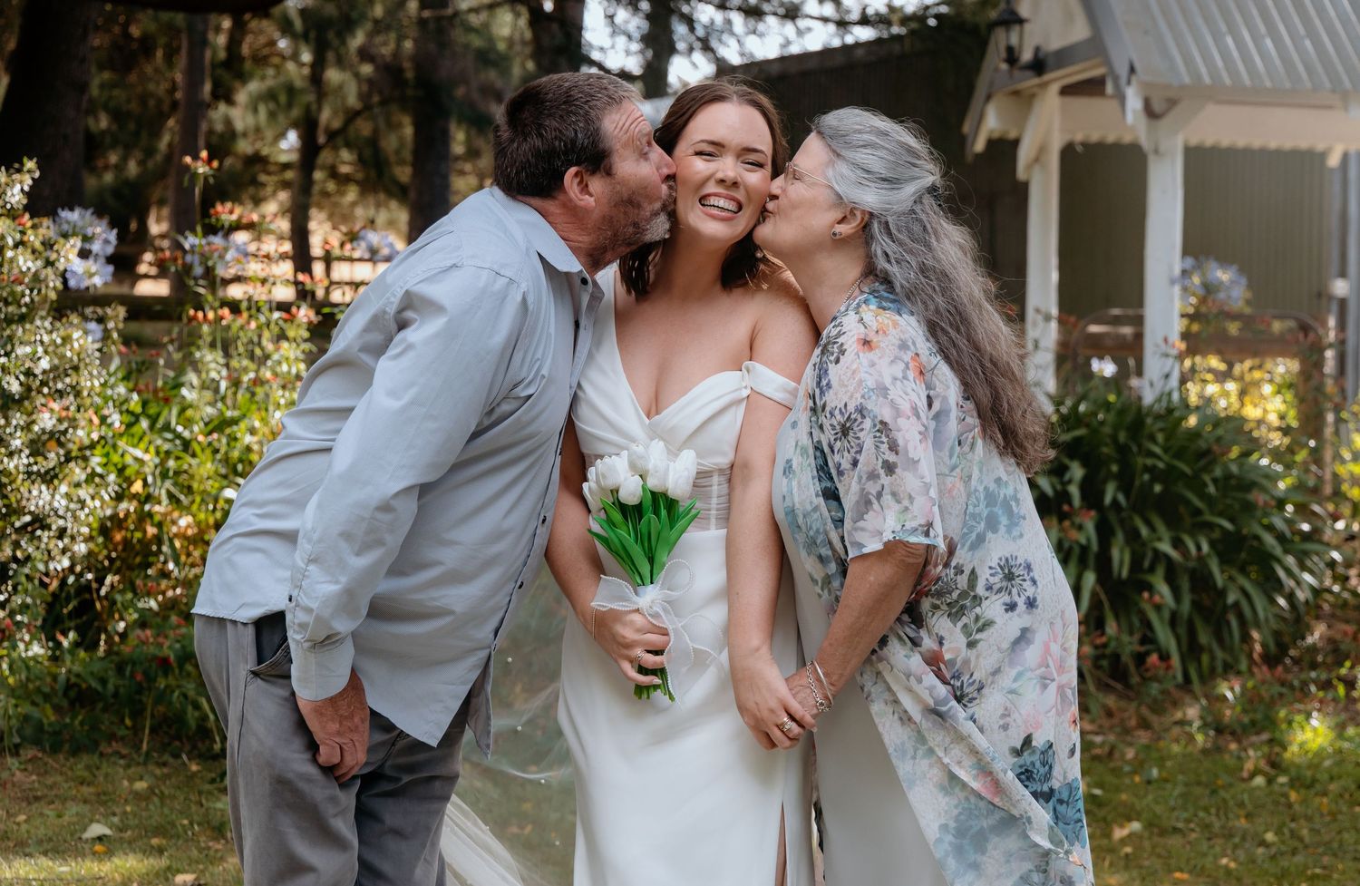 Bride in white gown holding bouquet receives kisses on cheeks from parents outdoors at wedding celebration.