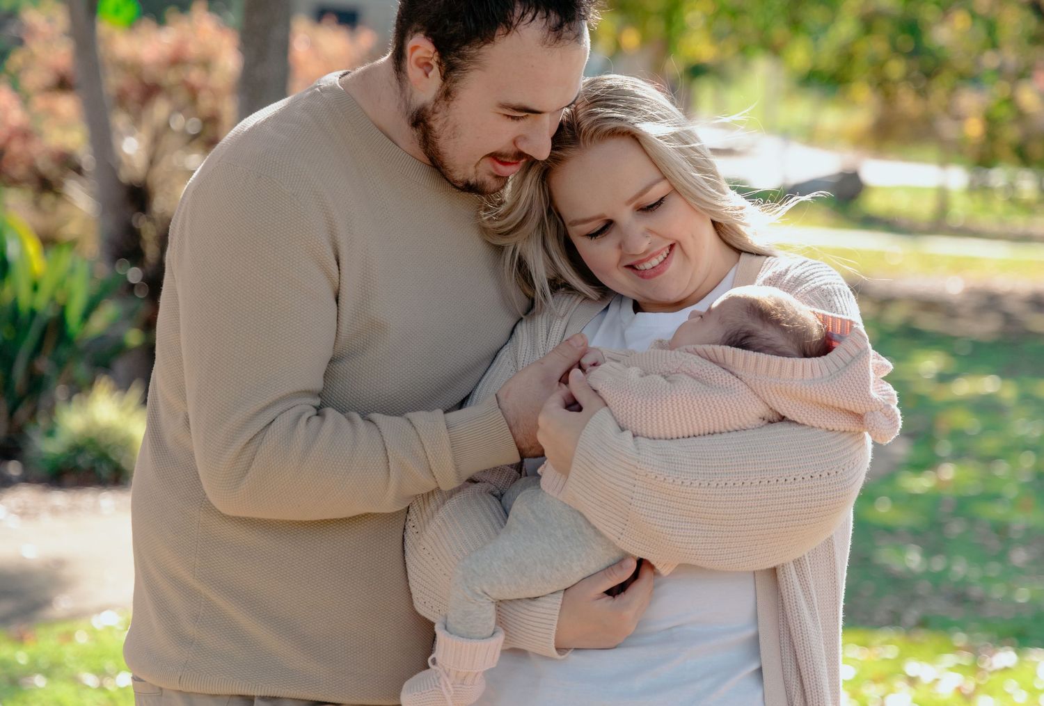 A couple in neutral toned clothing embraces tenderly in a sunlit outdoor setting with trees in the background.