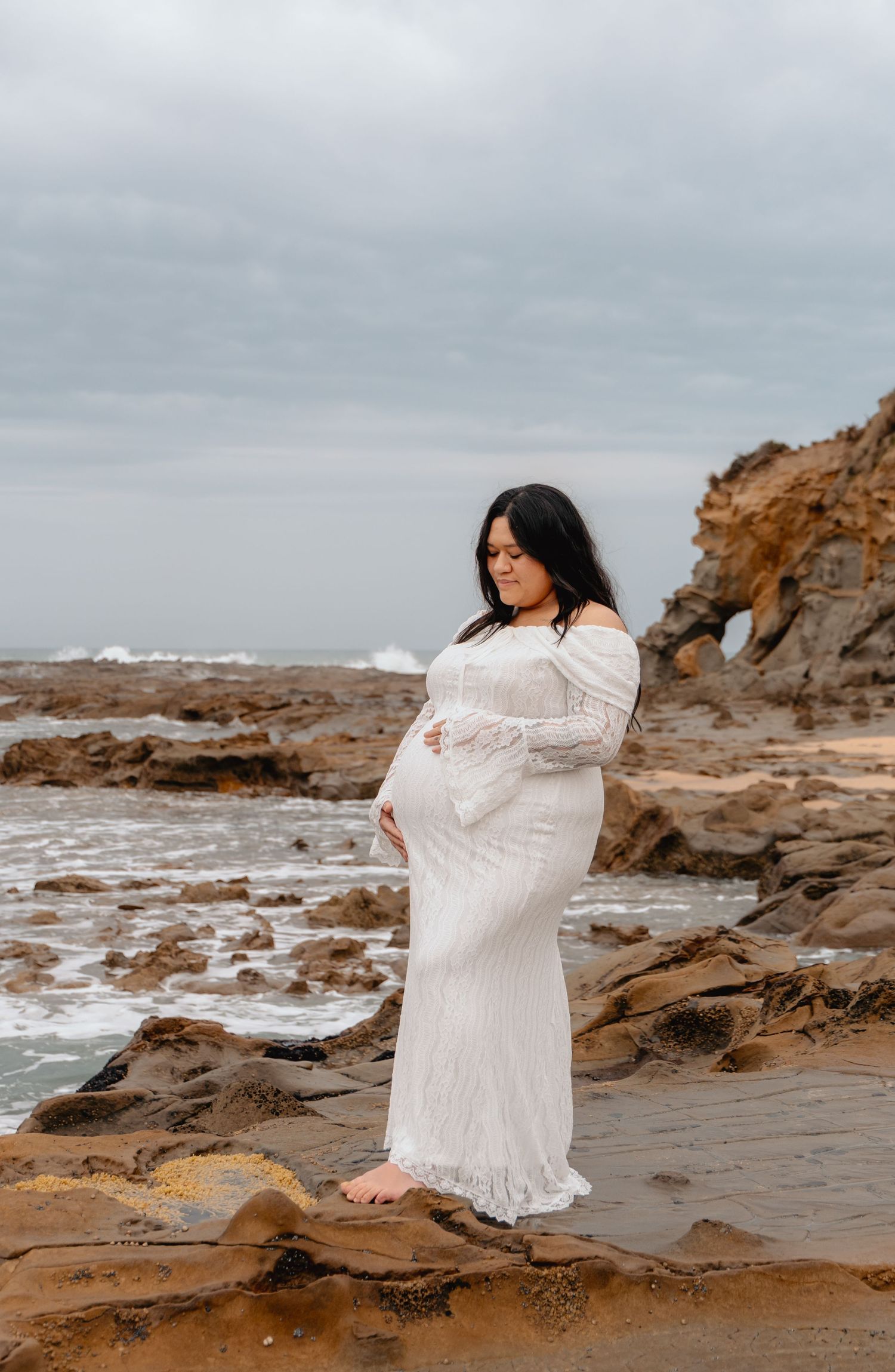 Expectant mother in white lace dress poses thoughtfully on rocky coastal shore during overcast day.