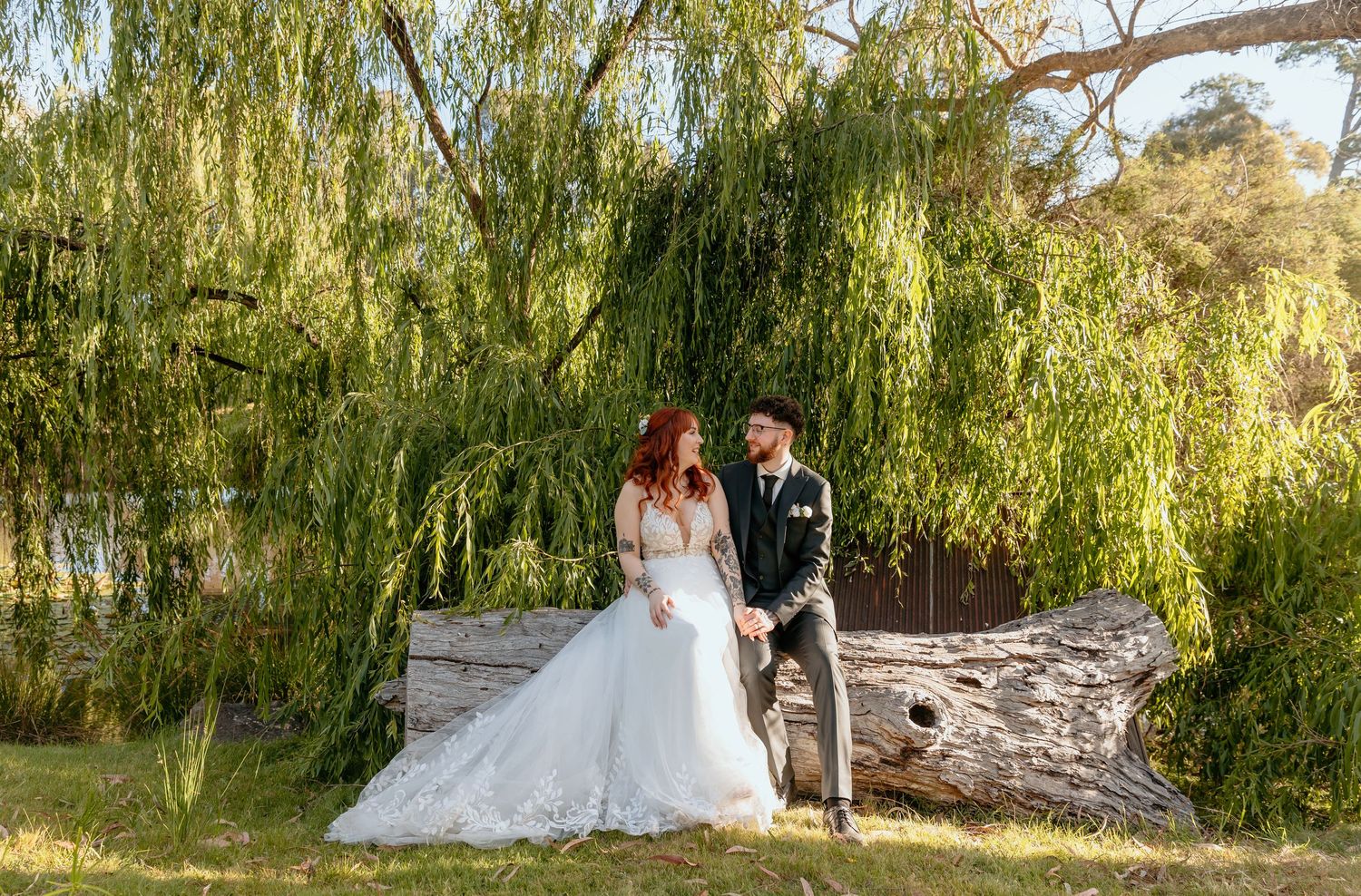 Wedding couple poses beneath cascading willow trees near a rustic stone bridge in a romantic garden setting.