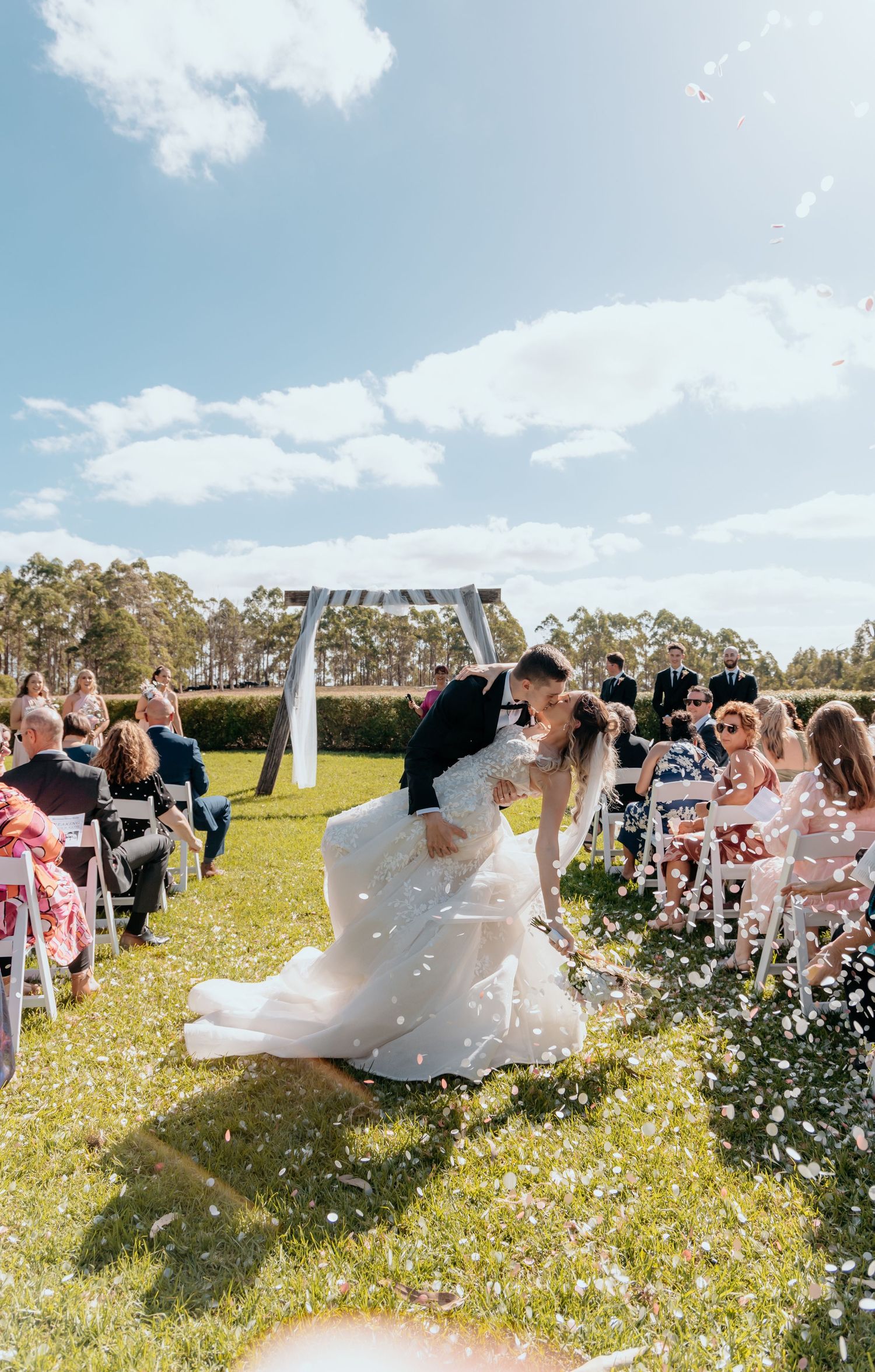 A couple celebrates at an outdoor wedding ceremony while guests shower them with white rose petals on a sunny day.