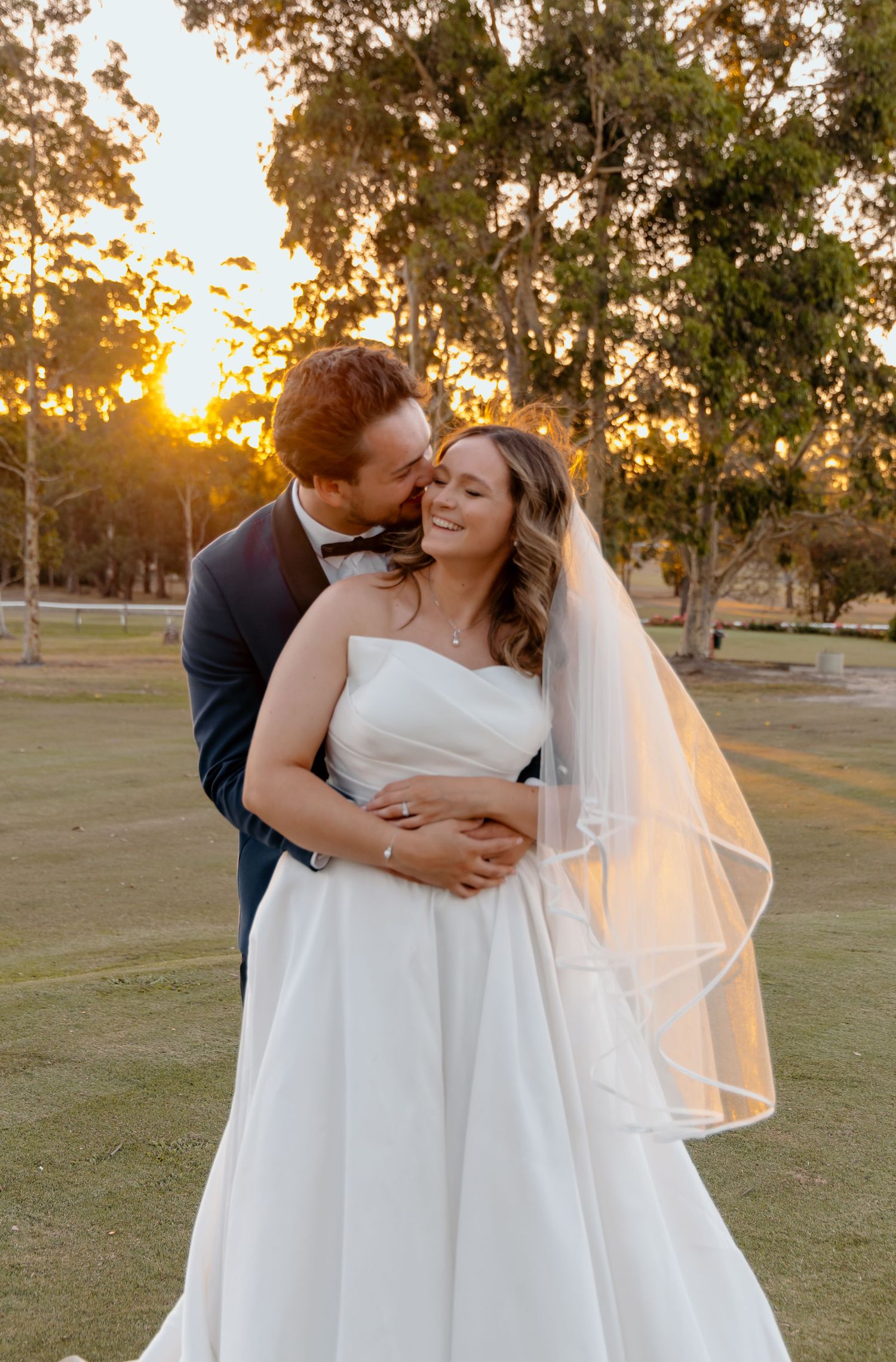 Romantic wedding couple embraces at sunset in an outdoor field with golden sunlight streaming through trees.