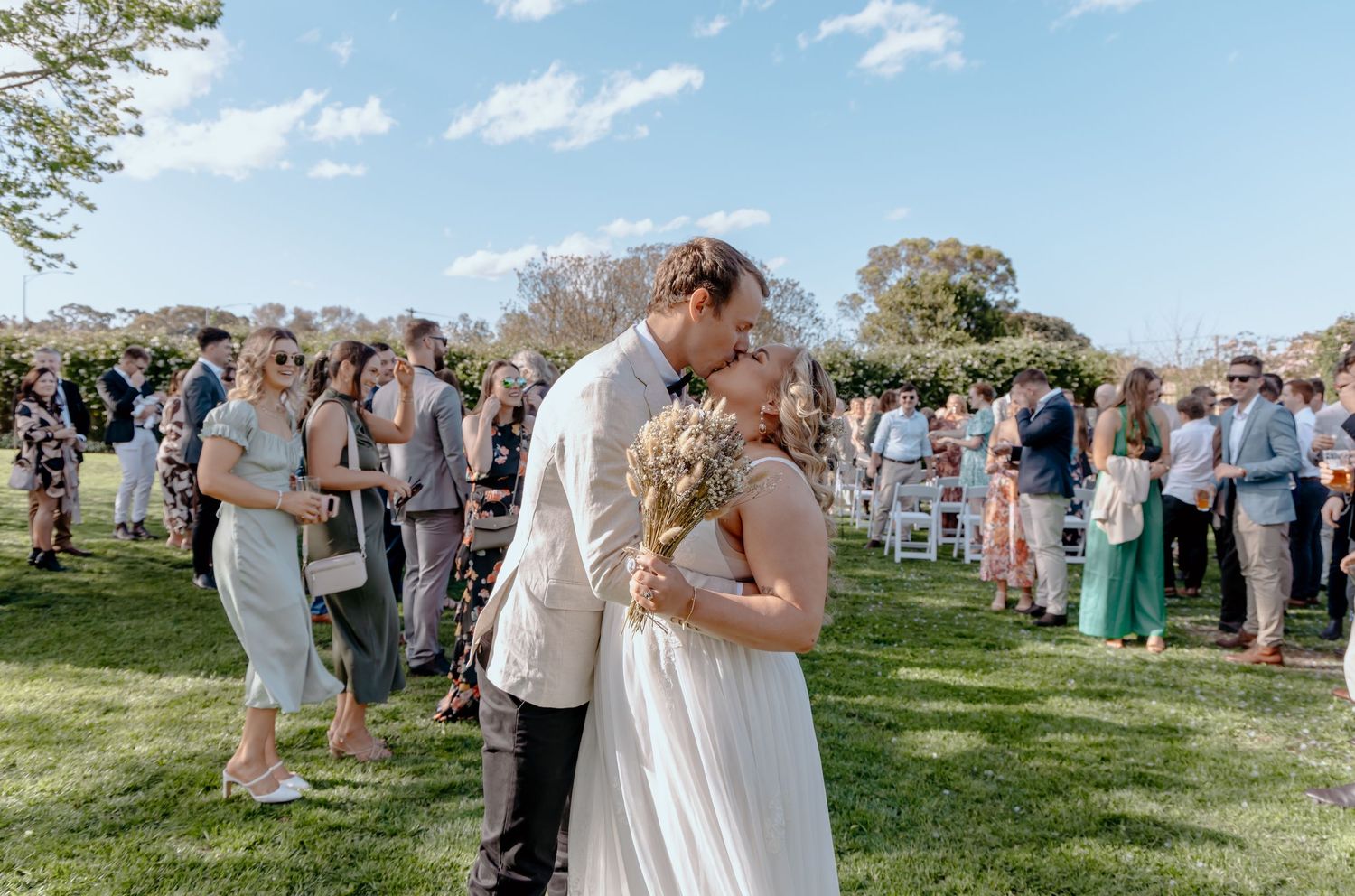 A couple shares a romantic kiss during their outdoor wedding ceremony while guests look on in the background.