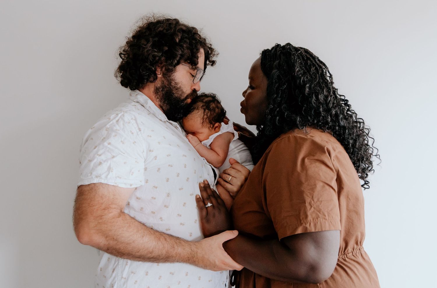 A couple shares a tender moment with their baby against a white wall.