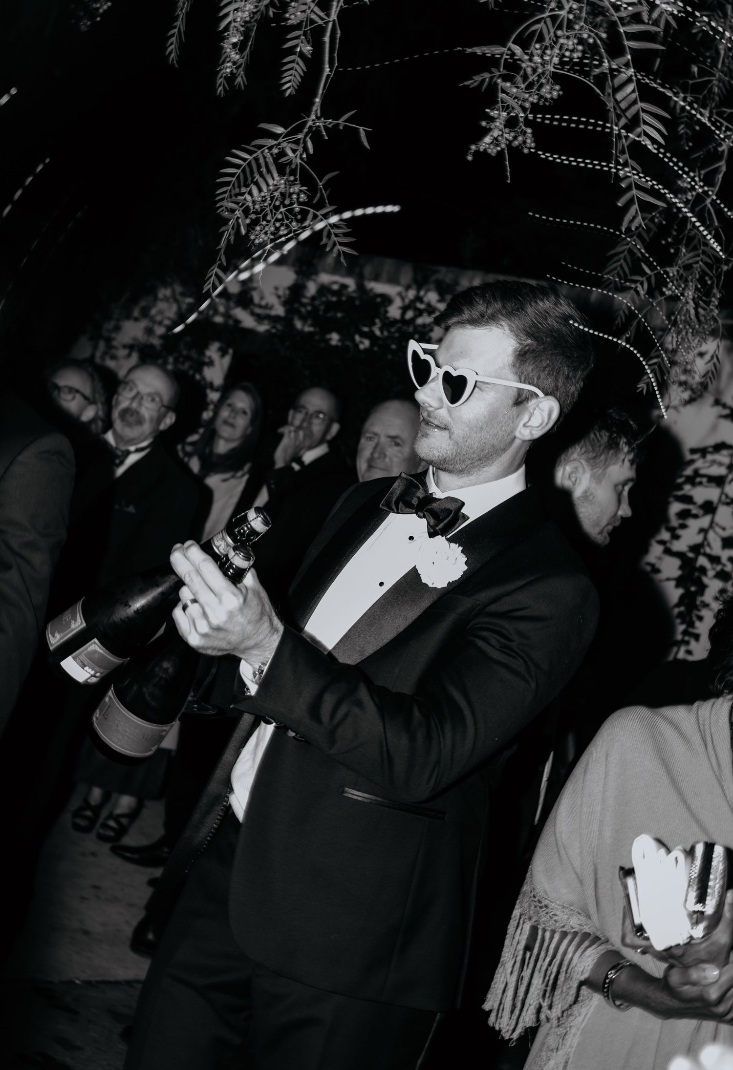 Man in tuxedo wearing heart-shaped sunglasses holding a champagne bottle at a wedding reception.