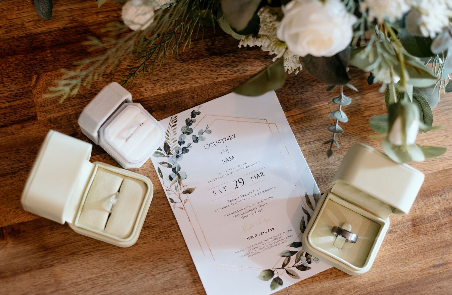 Elegant floral wedding invitations and vintage velvet ring boxes arranged with white roses and greenery on wooden surface.