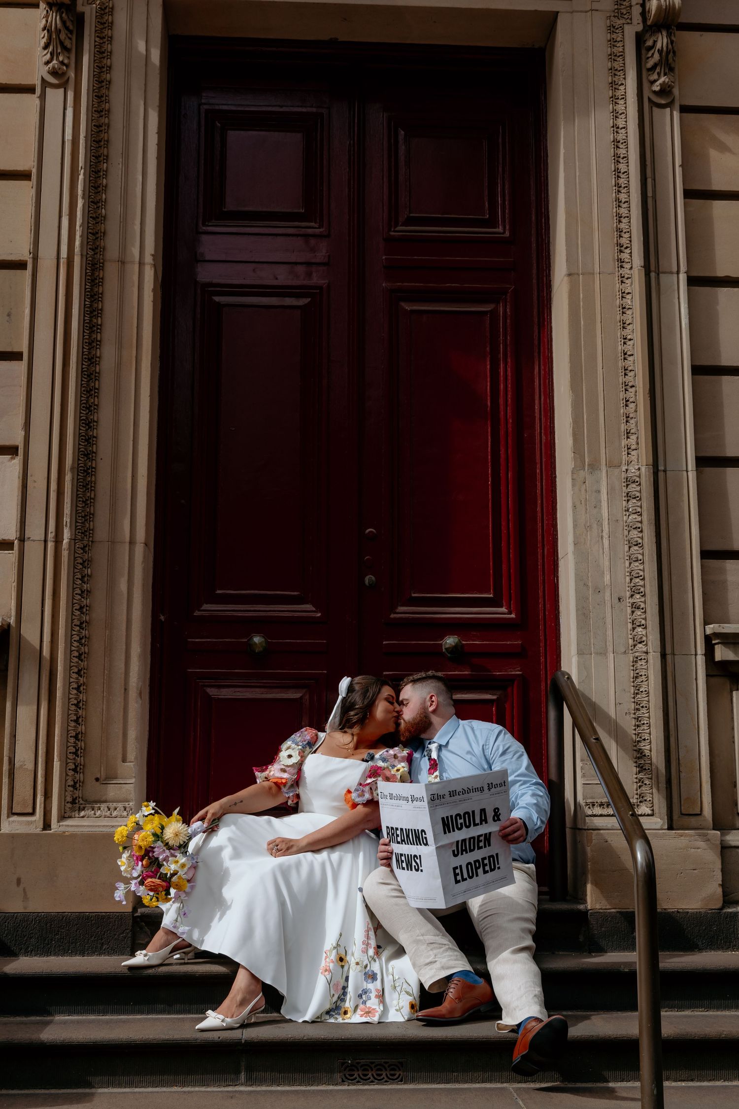 A couple sits on stone steps reading newspapers in front of a grand wooden door, with a bouquet of yellow flowers nearby.