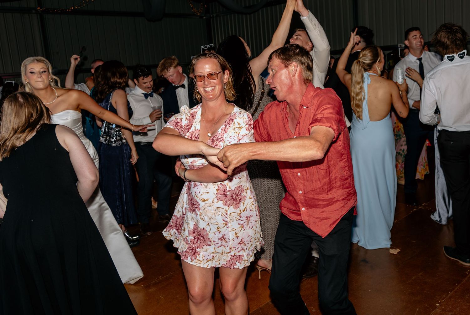 Couple dancing joyfully at a wedding reception on a crowded dance floor, woman in floral dress, man in red shirt.