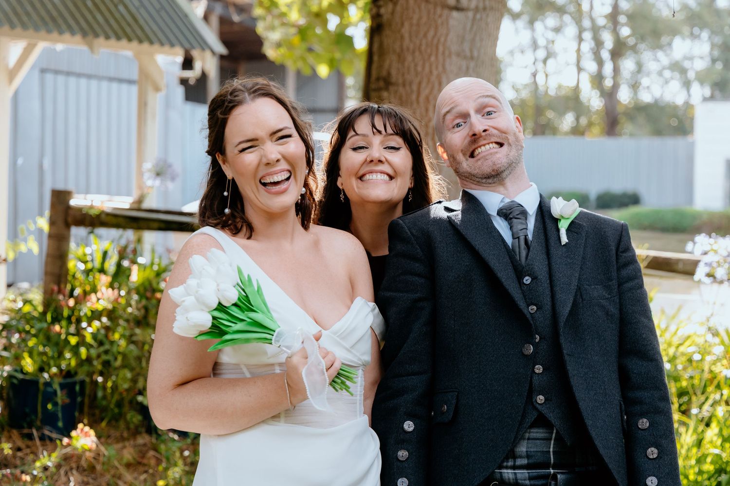 Joyful wedding trio with bride in white dress holding tulips, groom in tartan kilt, and celebrant smiling outdoors.