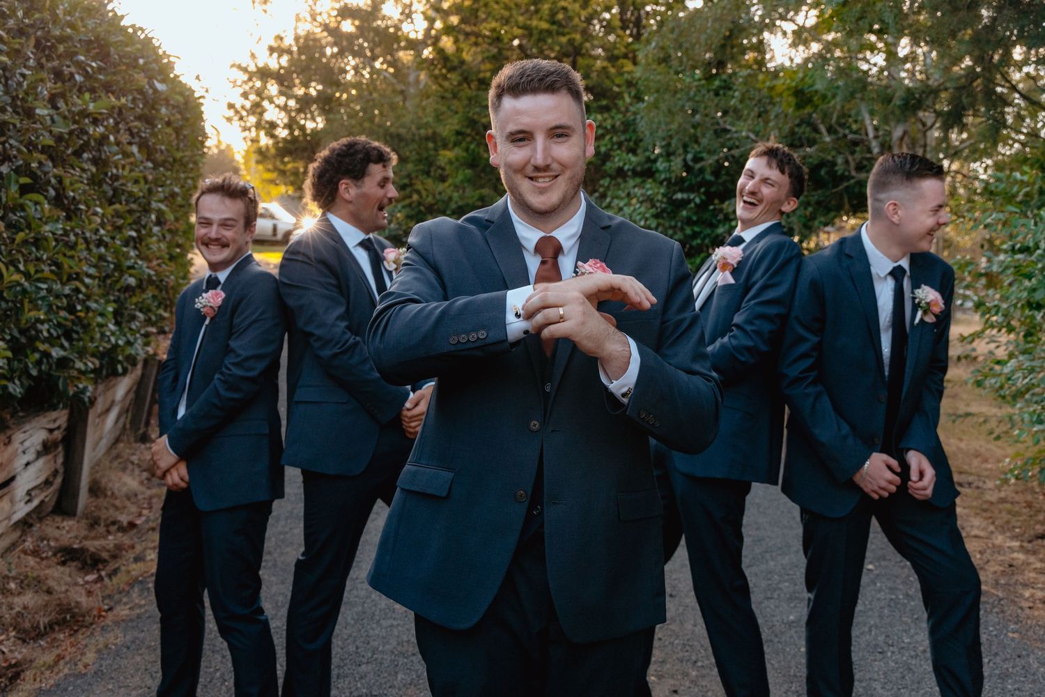 Groom adjusting cufflinks with smiling groomsmen in navy suits posing playfully behind him outdoors.