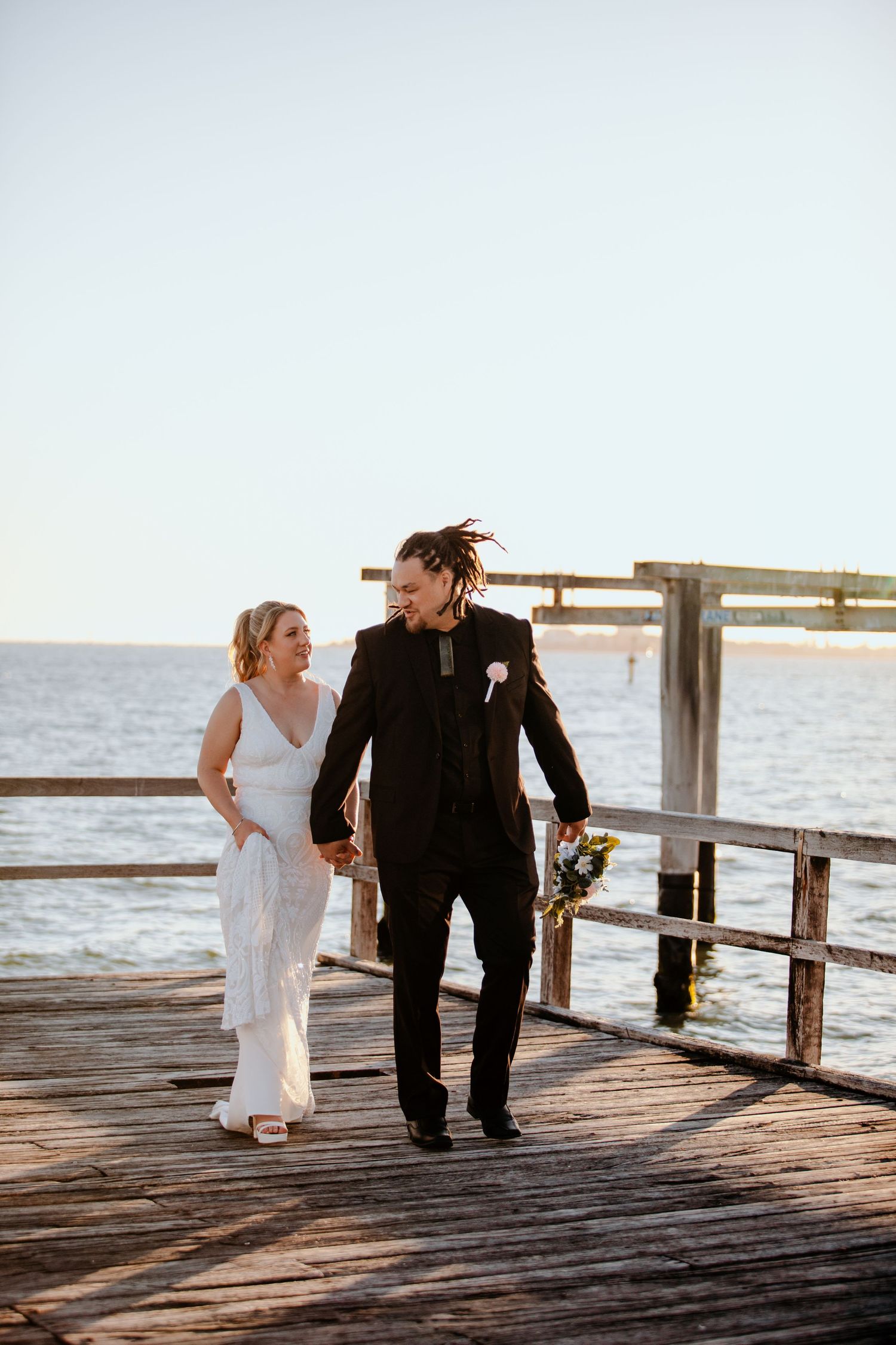 Newlywed couple walking hand-in-hand on a wooden pier by the ocean, bride in white dress, groom in black suit.