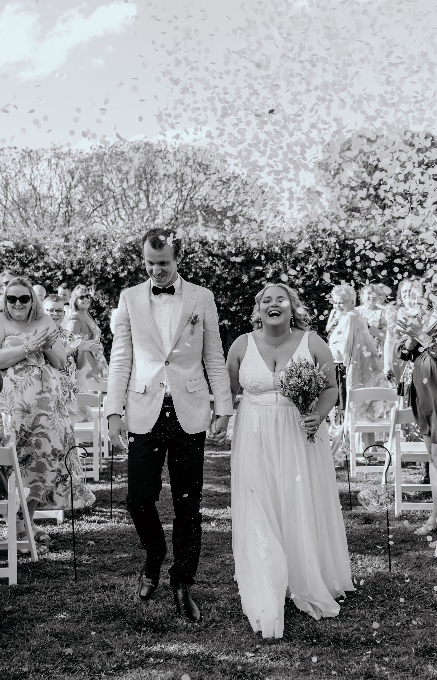 Wedding party walks down aisle through blooming apple orchard on sunny spring day.