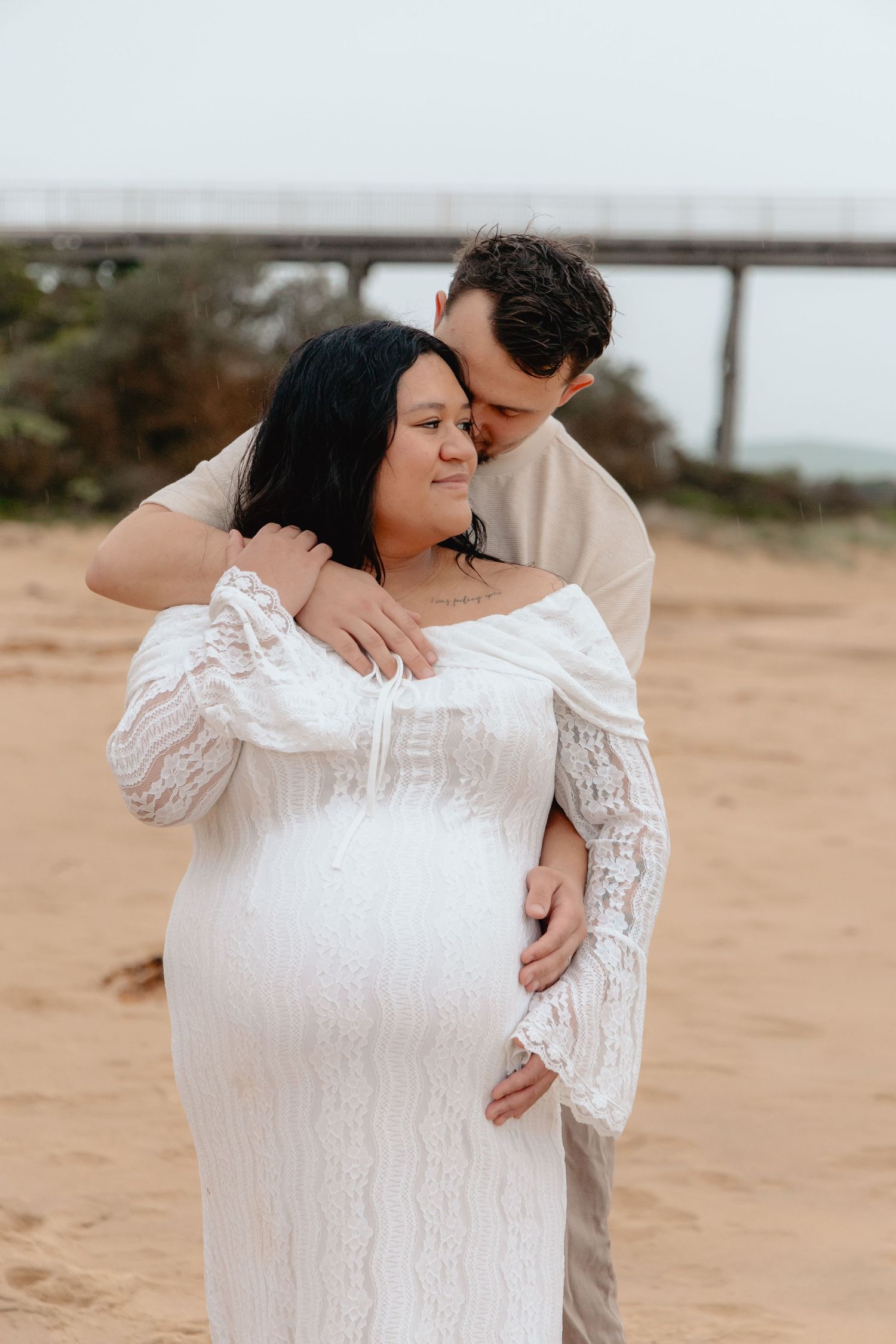 Intimate maternity photo on sandy beach with couple embracing near bridge in background.