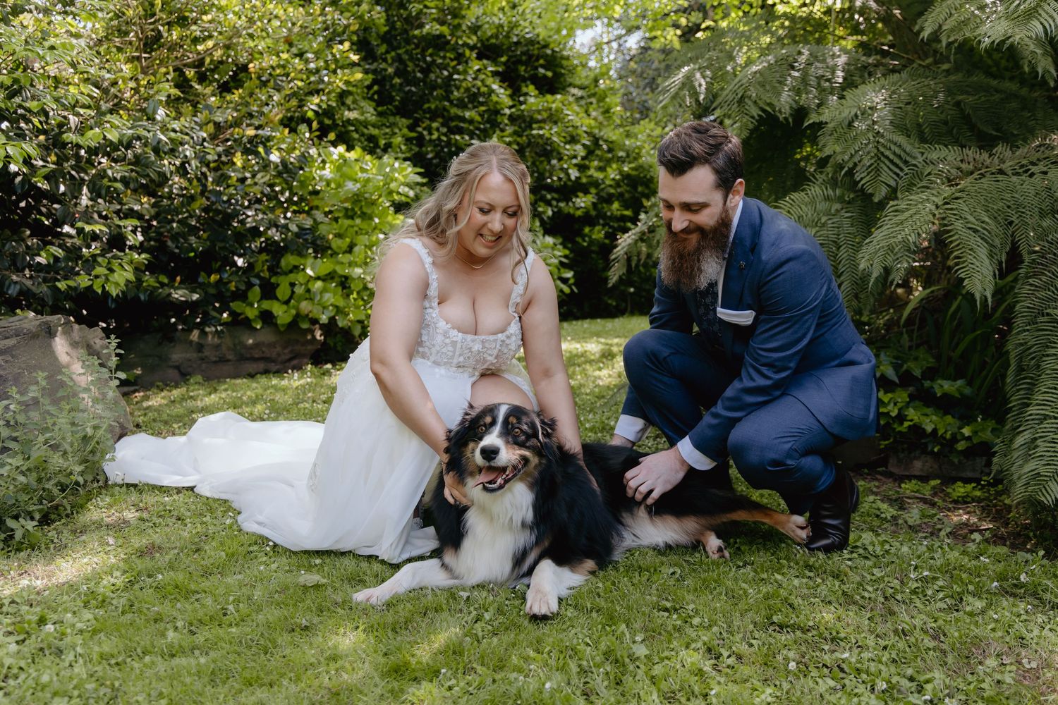 A couple in wedding attire sits on grass with their black dog in a lush garden setting.