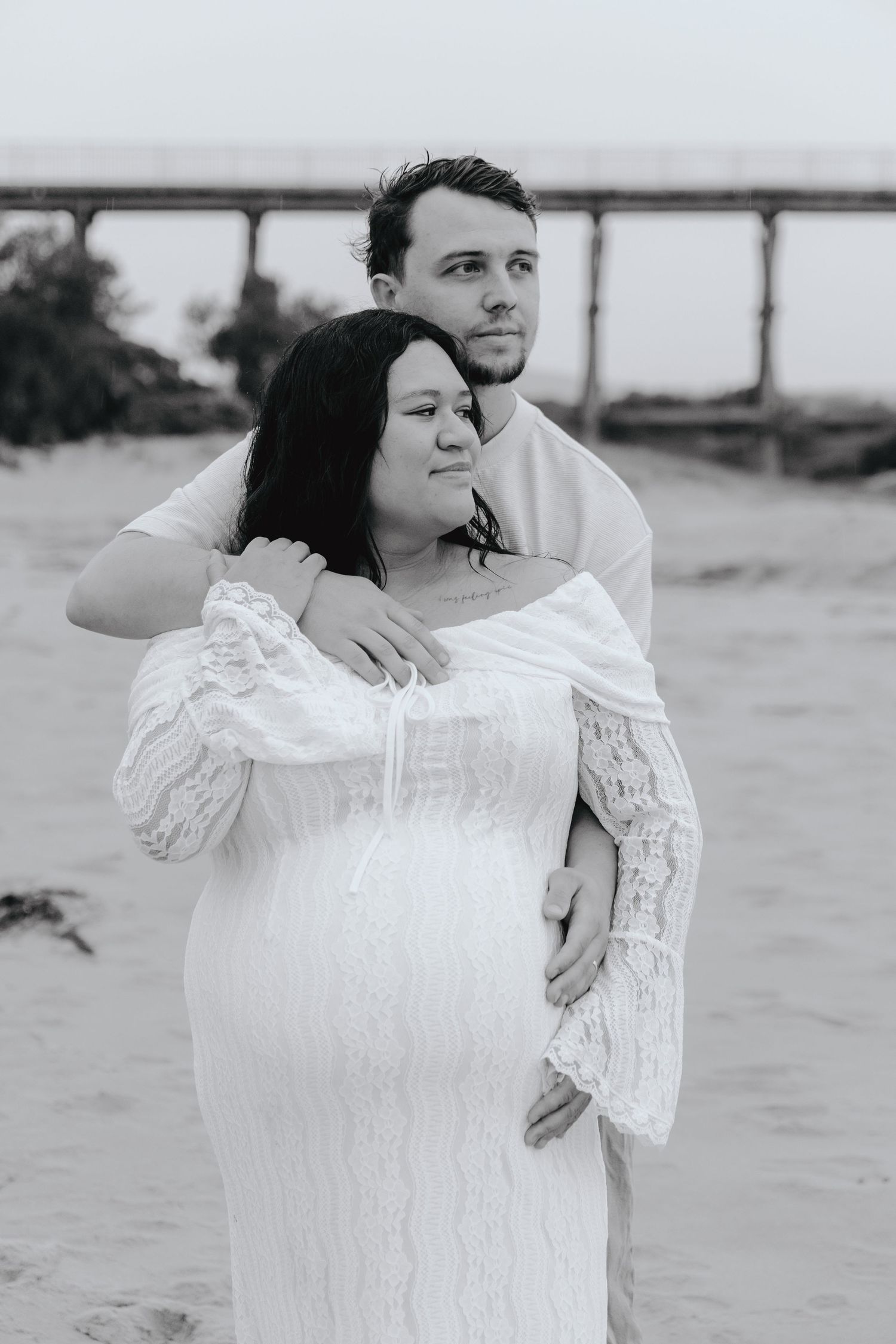 A black and white photo captures a couple dancing together on a beach with a bridge visible in the background.