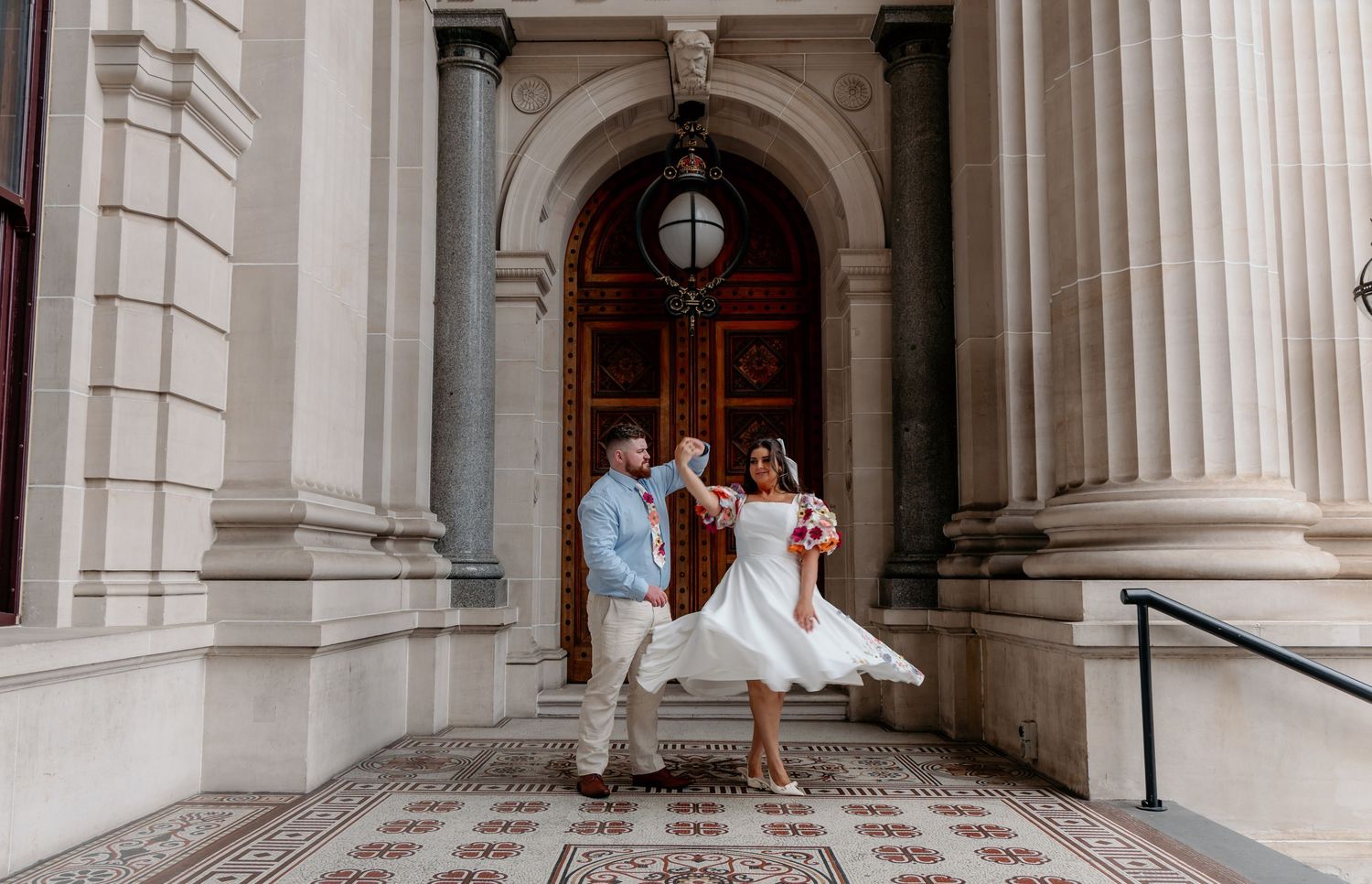 A couple in light summer attire poses playfully in front of an elegant arched doorway with classical stone columns.