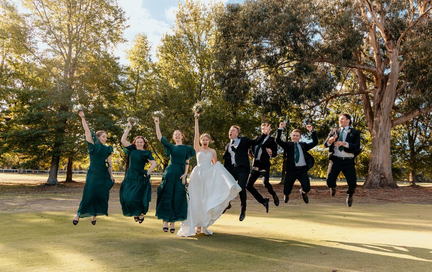 A joyful wedding party in green dresses and suits leap into the air together in a scenic outdoor setting.