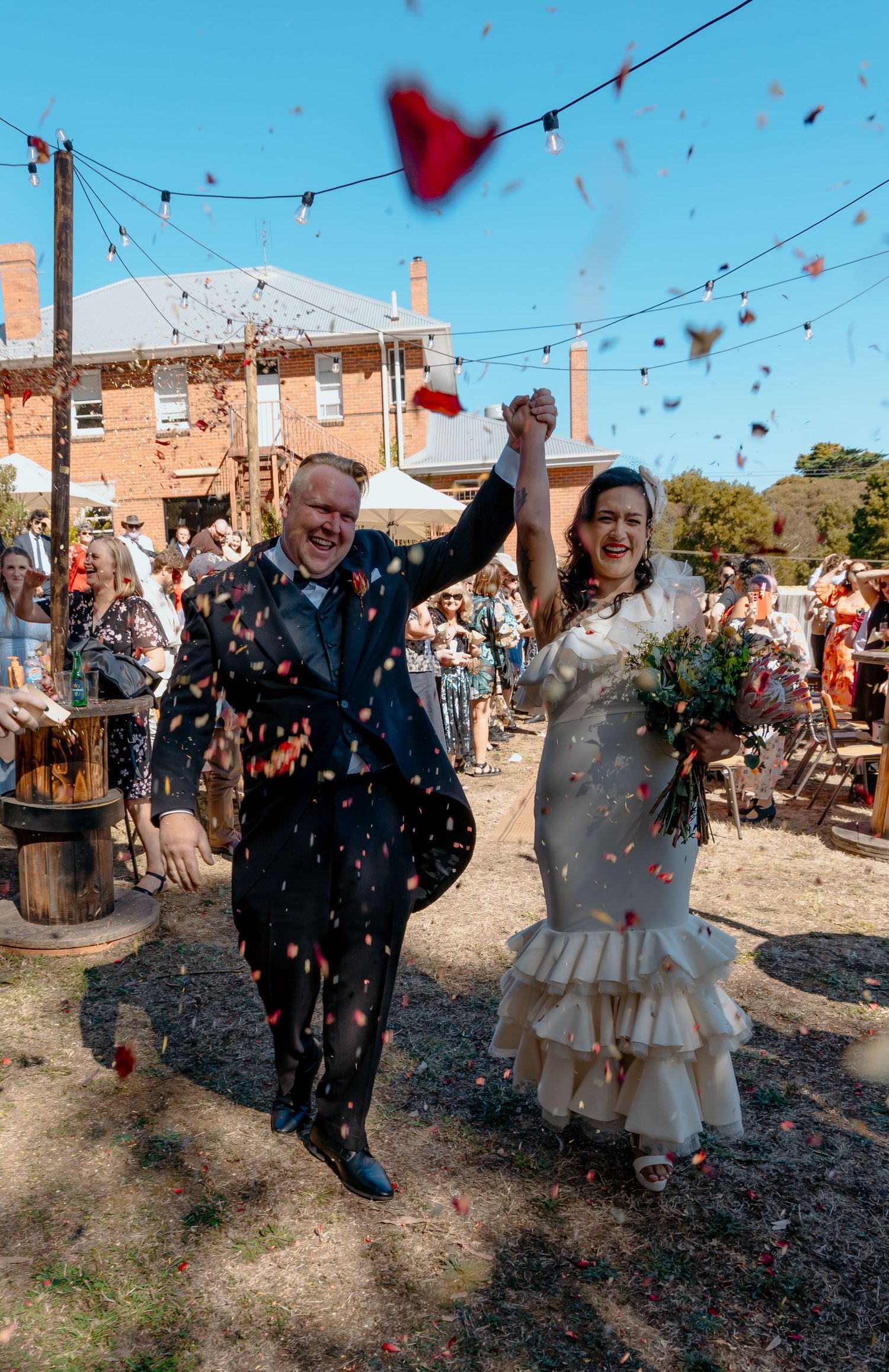 Couple celebrating at outdoor wedding venue with string lights and red confetti floating through the air.