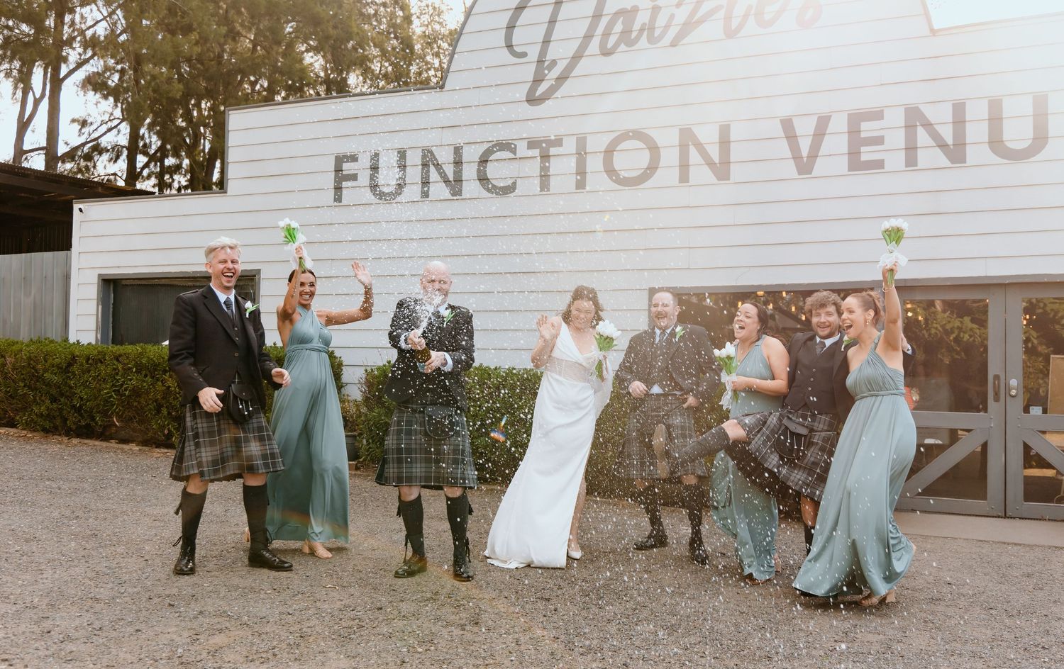 Wedding party celebrates outside a Scottish-themed function venue, with kilts, sage bridesmaid dresses, and champagne.