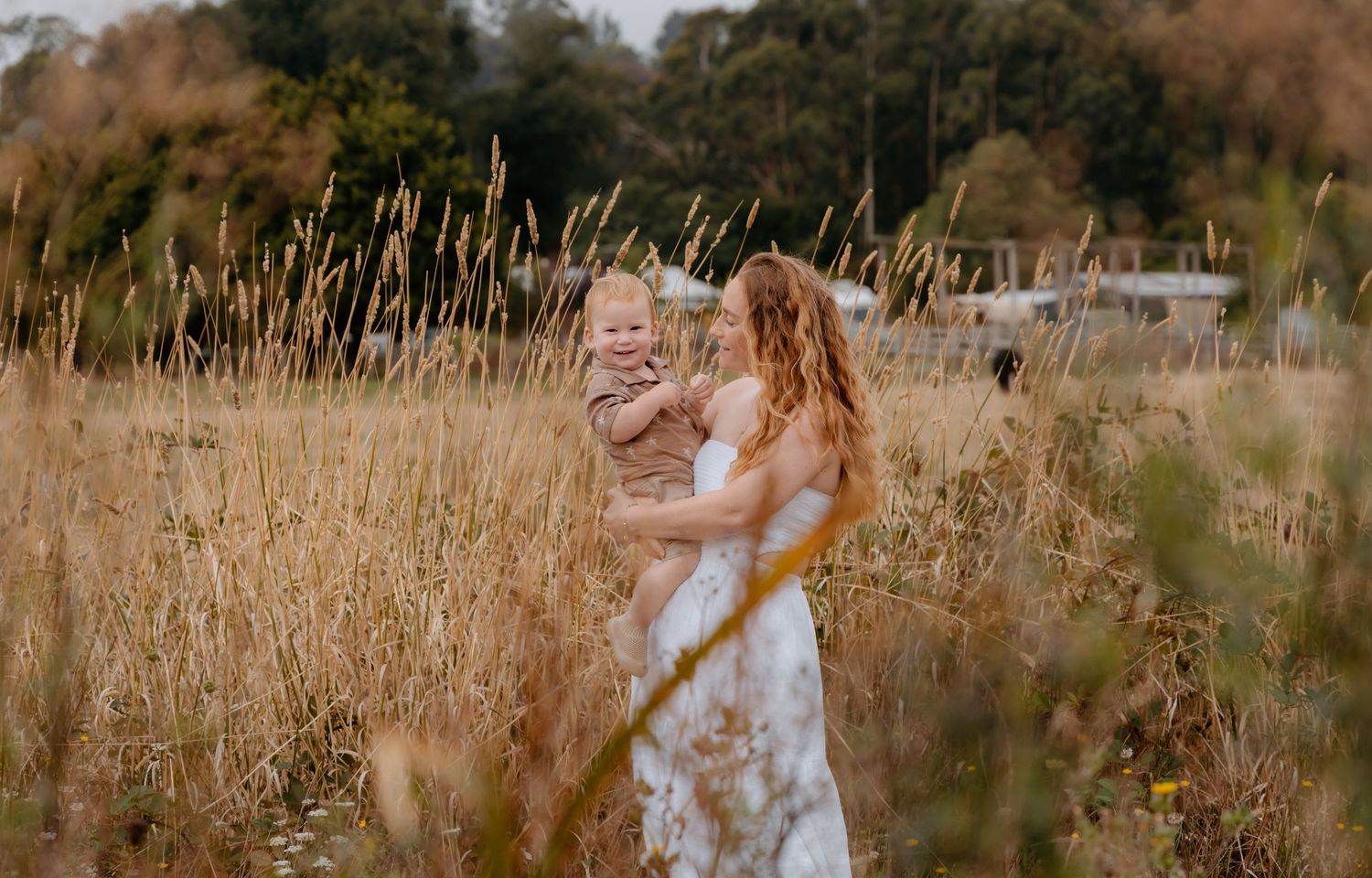 A mother in a floral dress holds her baby in a golden field of tall grass outdoors.