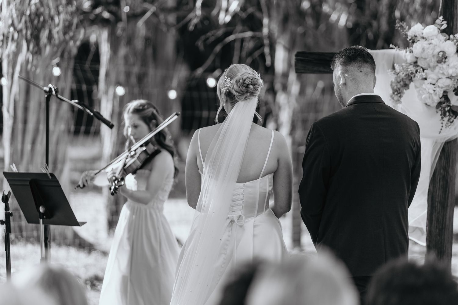 Black and white wedding ceremony with bride and groom at altar, violinist performing, floral arch backdrop.